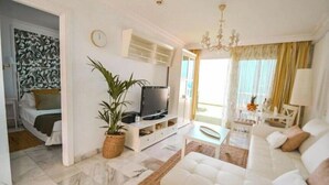 A modern living room with a white sofa, a TV on a white stand, a potted plant, and a chandelier. - Apartment - LOS GIGANTES (Santiago del Teide)