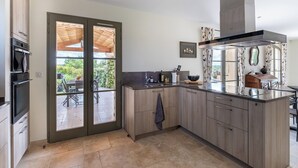 A modern kitchen with a large island, wooden cabinets, and a sliding glass door leading to an outdoor patio. - Spacious, comfortable stone farmhouse with swimming pool for 8 people in Uzès (GARRIGUES STE EULALIE)