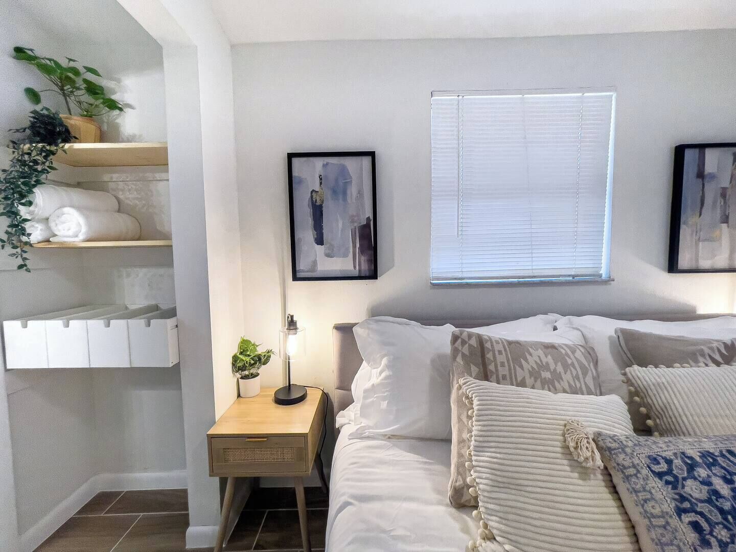 A bedroom with a bed, a nightstand, a window with blinds, and wall-mounted artwork.