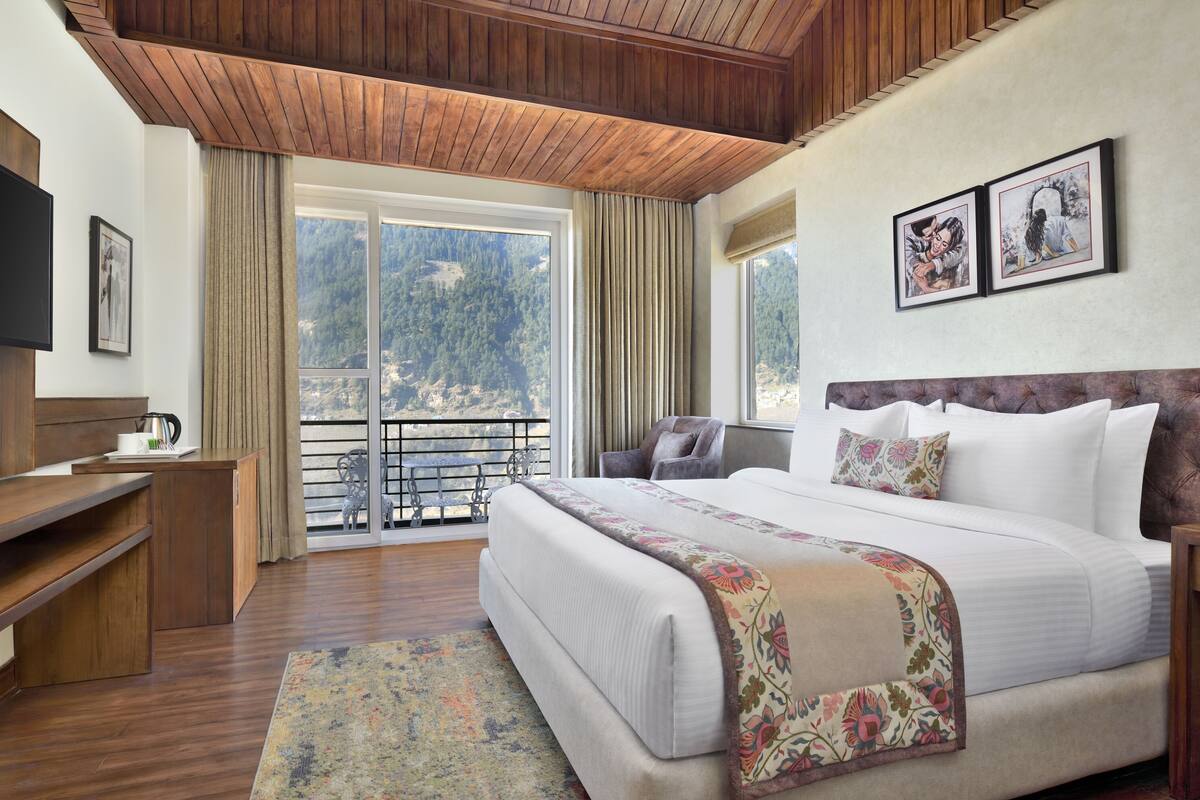 A hotel room with a large bed, a wooden desk, a television, and a balcony with a view of the mountains.