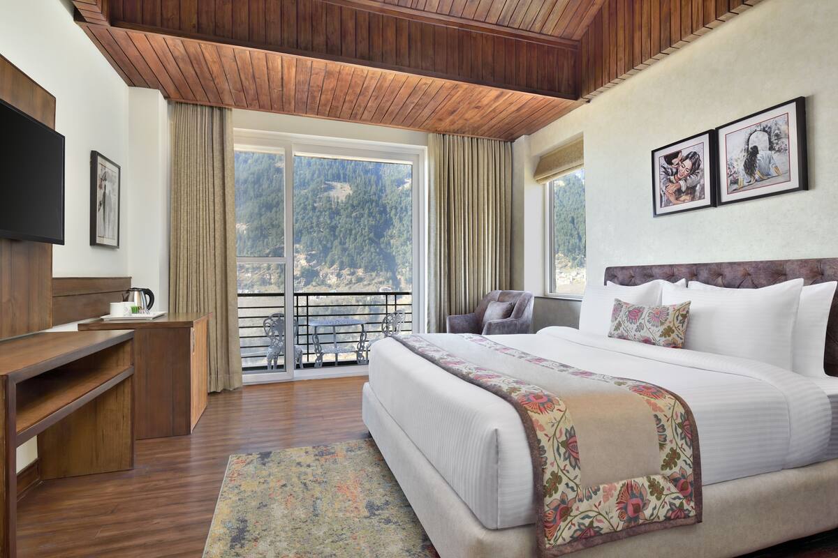 A hotel room with a bed, a desk, a chair, a TV, and a balcony with a view of the mountains.