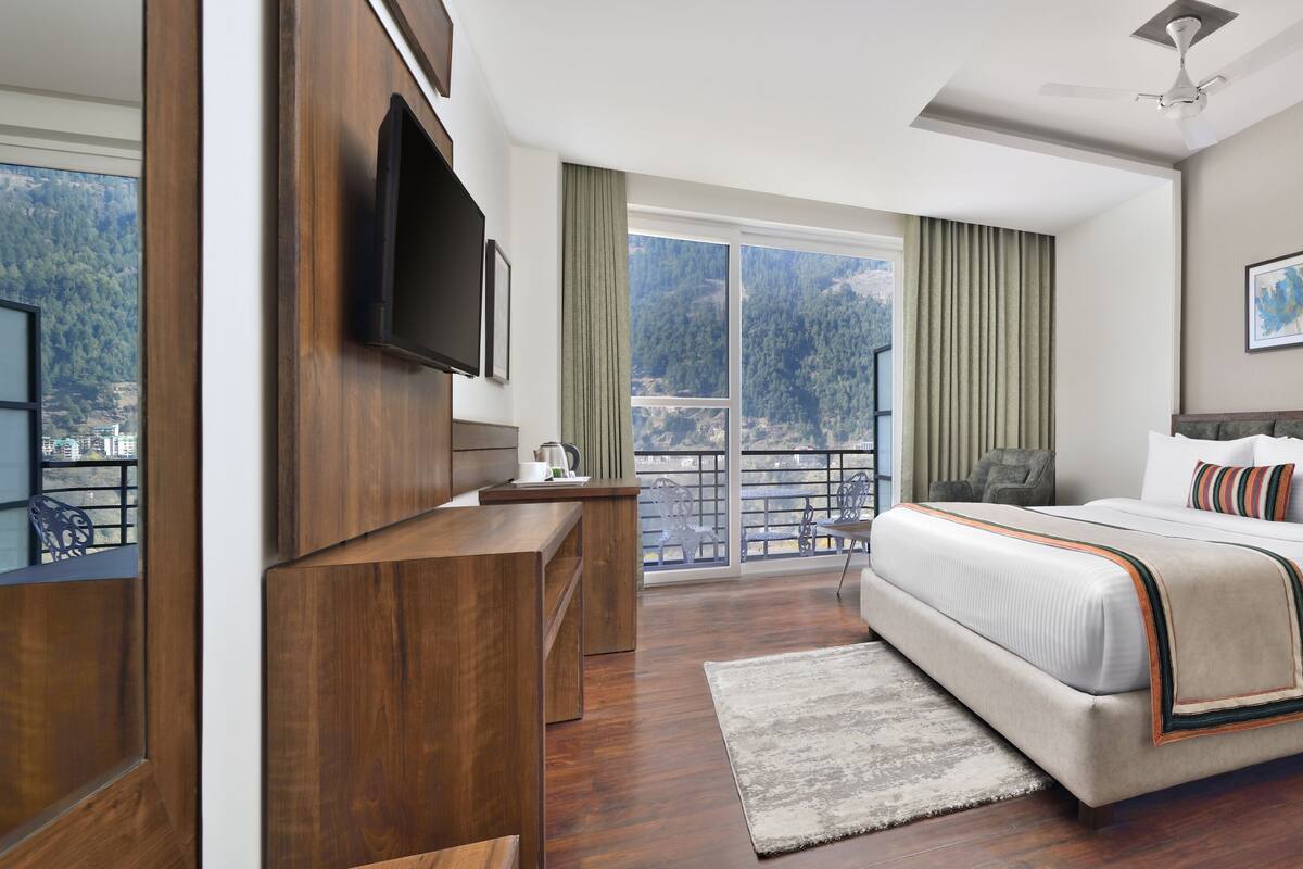 A hotel room with a large bed, a desk, a TV, and a balcony with a view of the mountains.