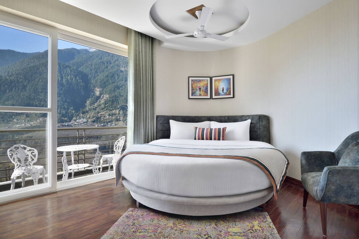 A bedroom with a large bed, a ceiling fan, a balcony with chairs, and a mountain view.