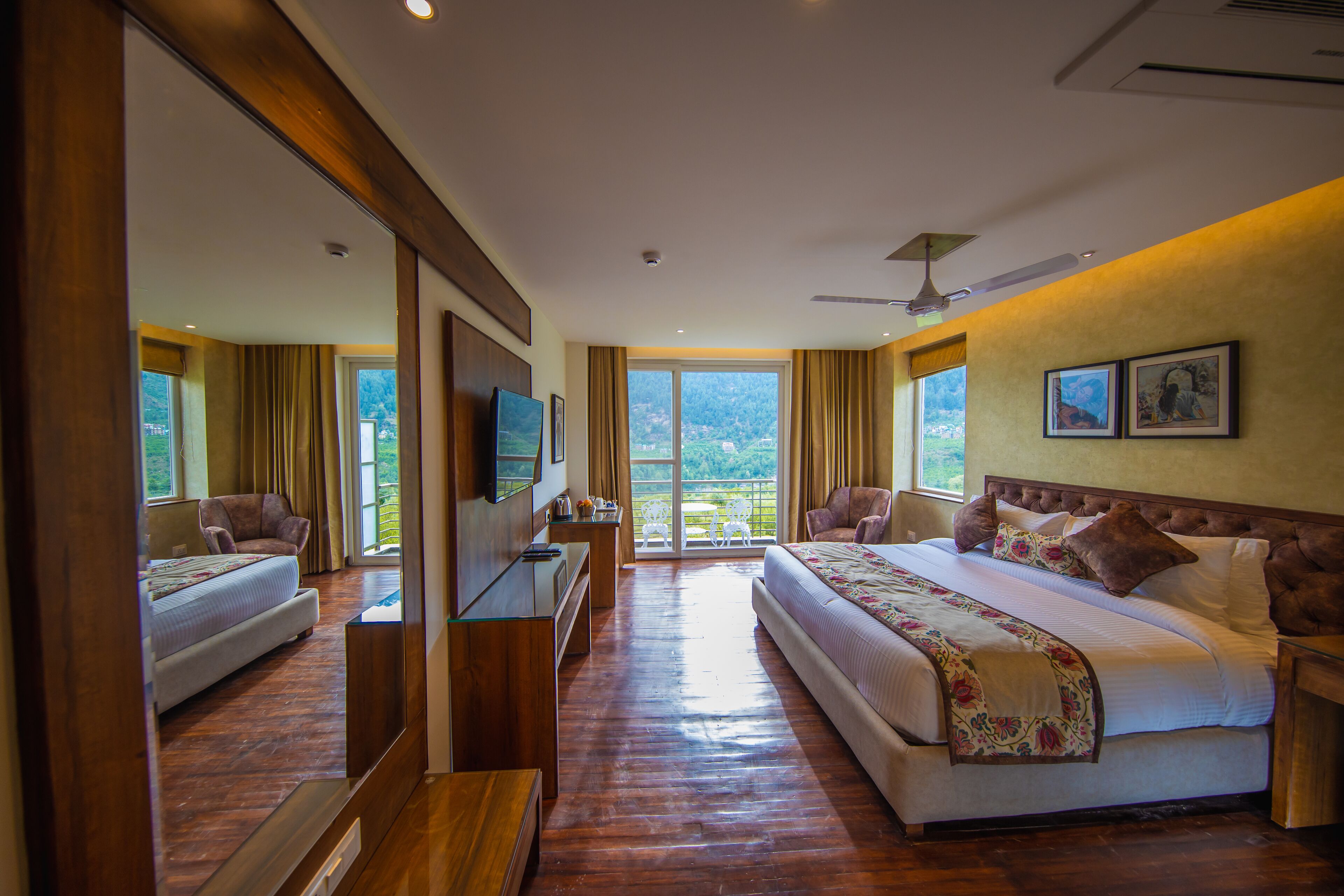 A spacious bedroom with a large bed, a desk, and a television. There is a balcony with a view of the mountains.