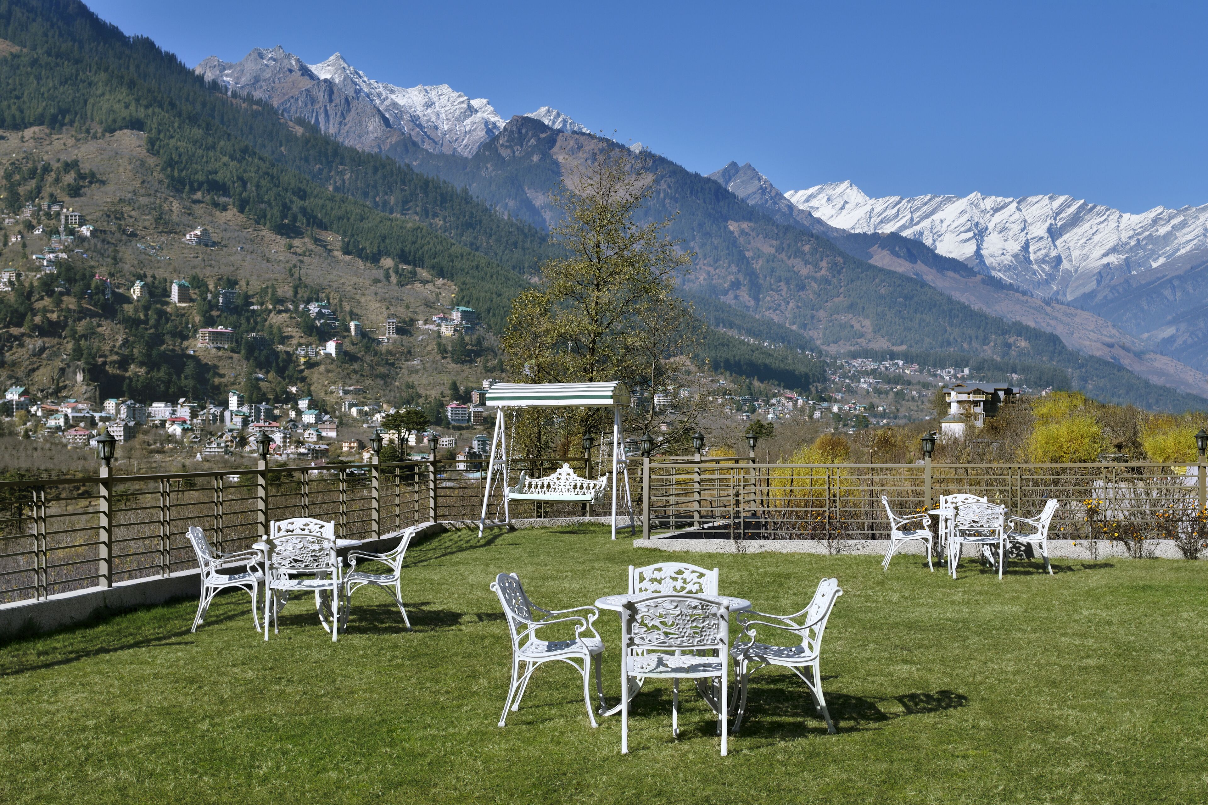 A terrace with white metal chairs and tables, overlooking a mountainous landscape with snow-capped peaks and a town below.