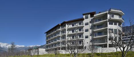 Un hotel de varios pisos con balcones, rodeado de un paisaje con césped y montañas al fondo.