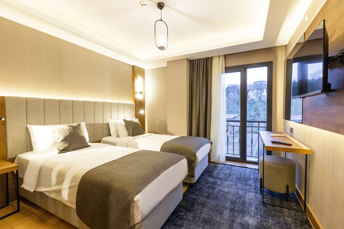 Habitación de hotel con dos camas, un escritorio, una silla, un televisor y un ventanal con vista a la ciudad.