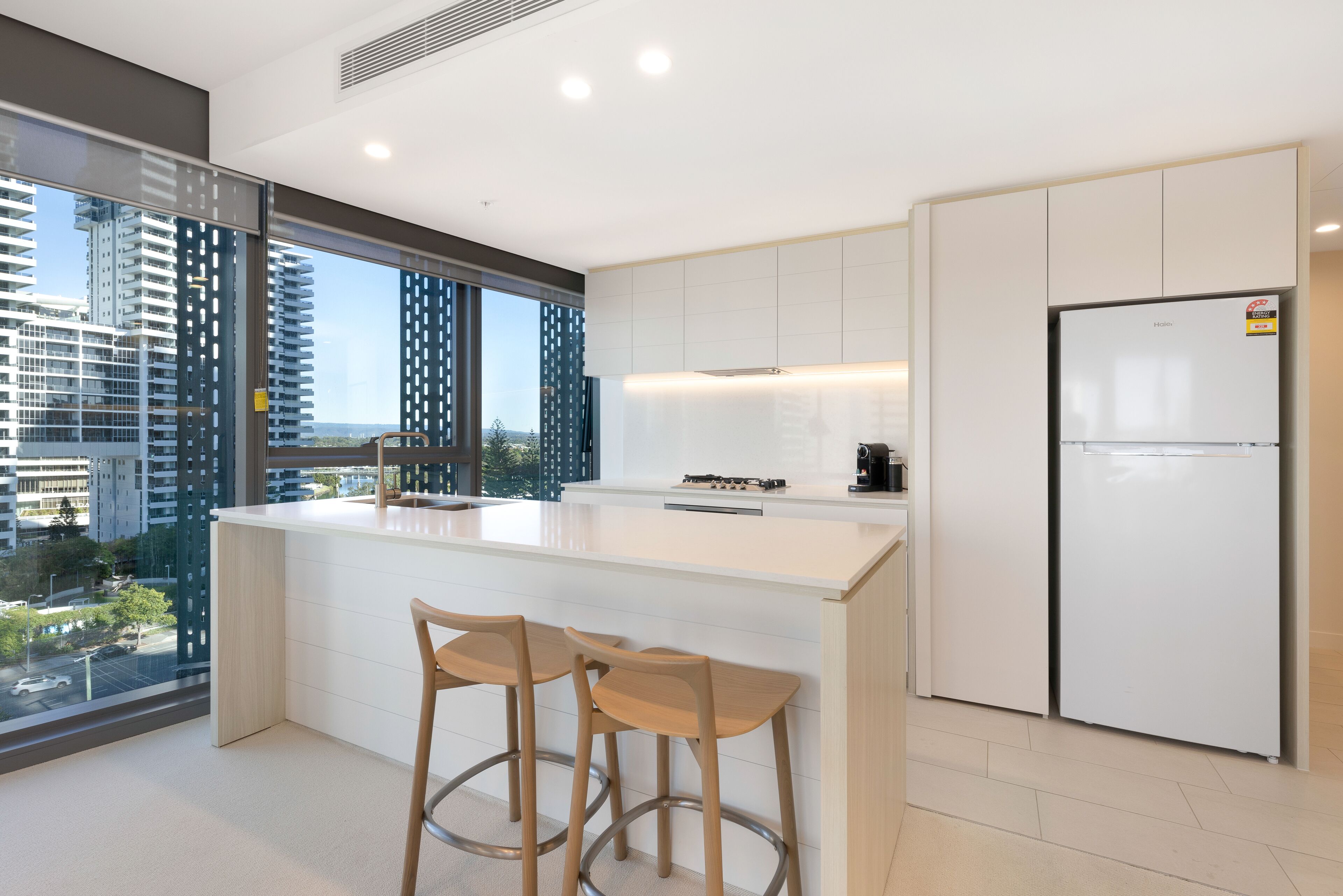 A modern kitchen with a large island, two bar stools, a built-in stove, and a refrigerator.
