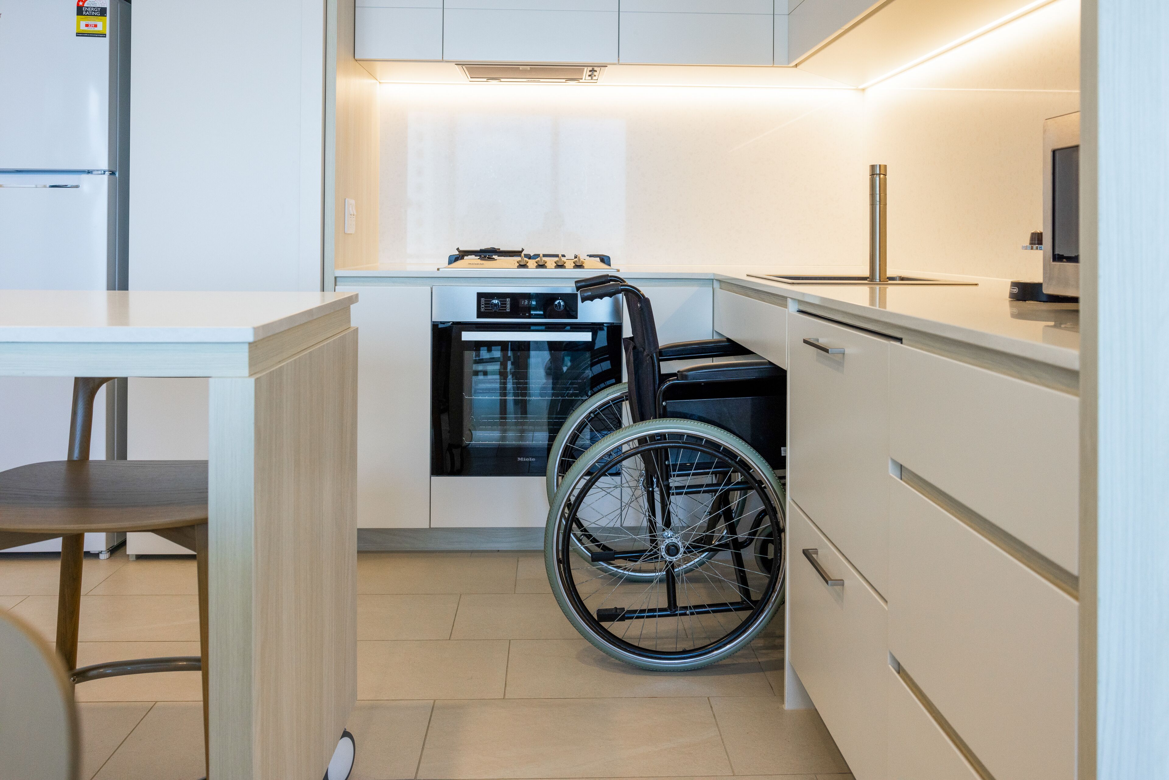 A modern kitchen with white cabinetry, a built-in oven, and a wheelchair in the corner.