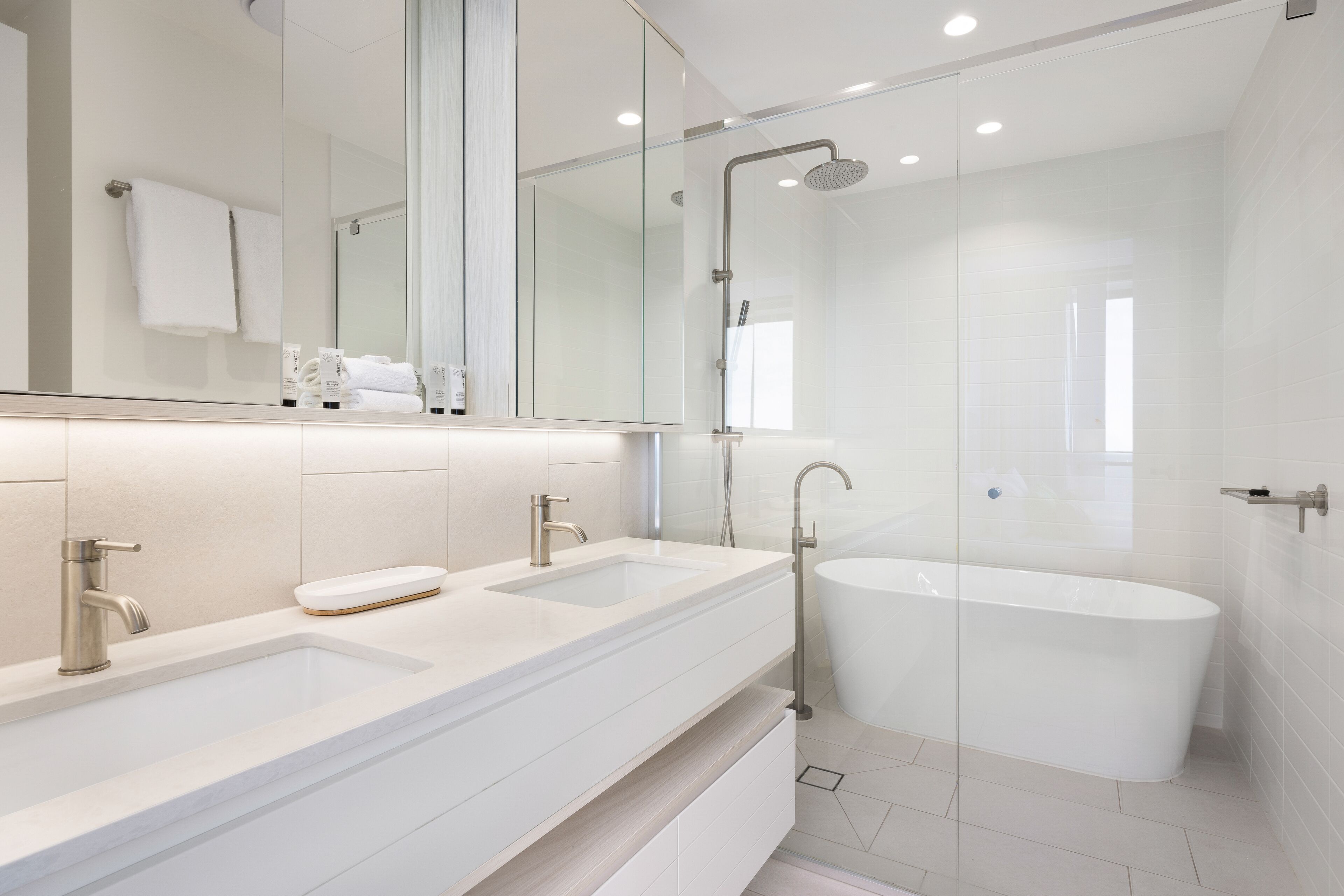 Un baño moderno con un espejo grande, dos lavamanos, una bañera y una zona de ducha con paredes de vidrio.