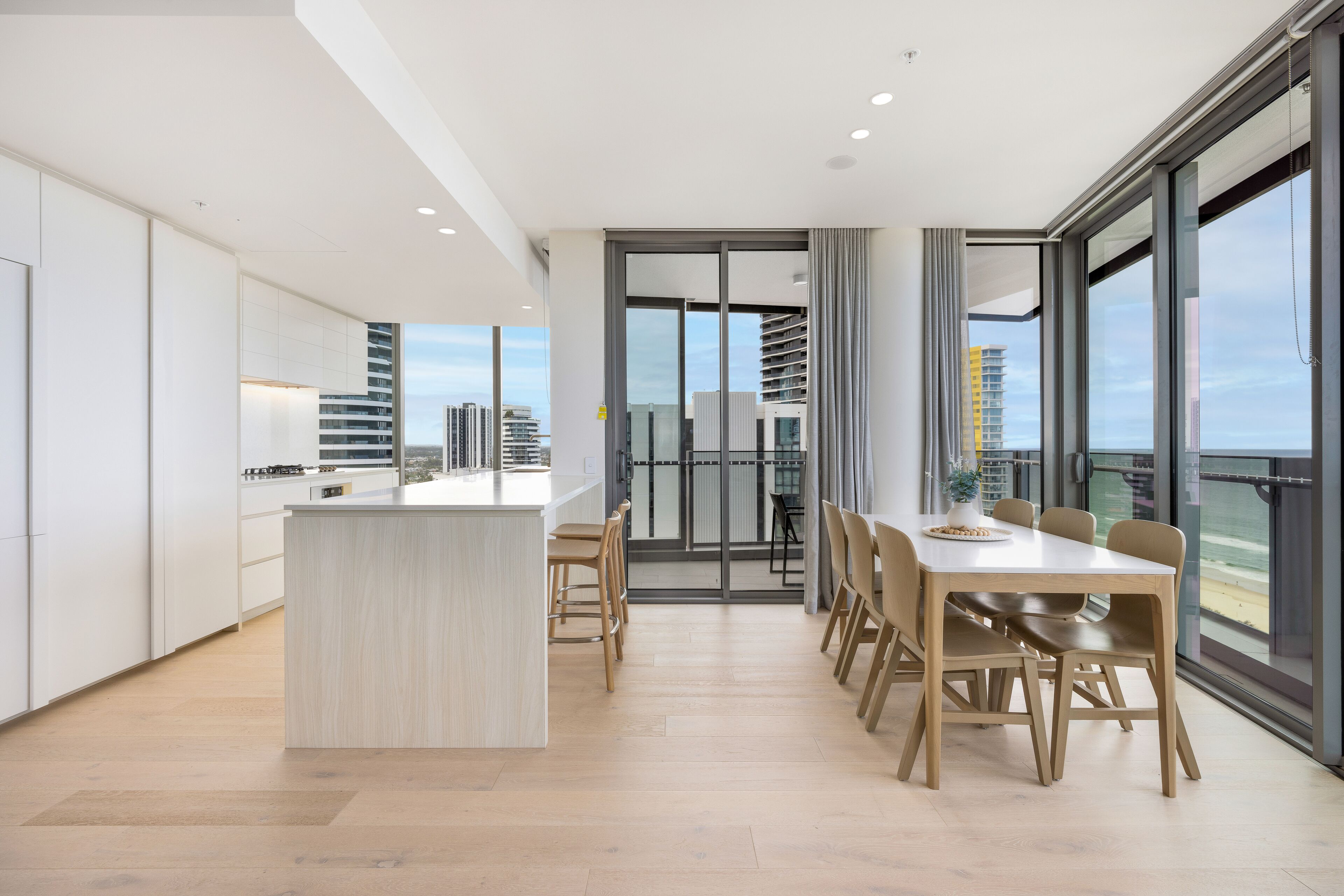 A modern kitchen with a central island, a dining area with a large table and chairs, and large windows offering a city view.