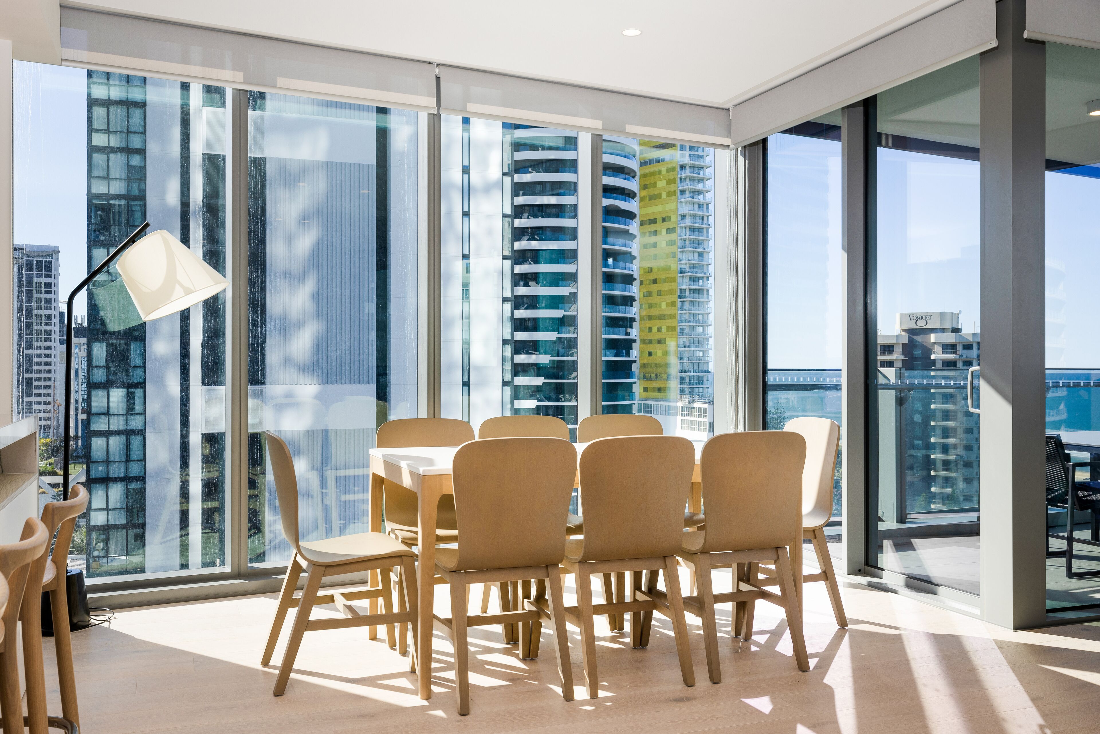 Un comedor moderno con una mesa grande y sillas, ventanas amplias con vista a la ciudad y una lámpara de pie.