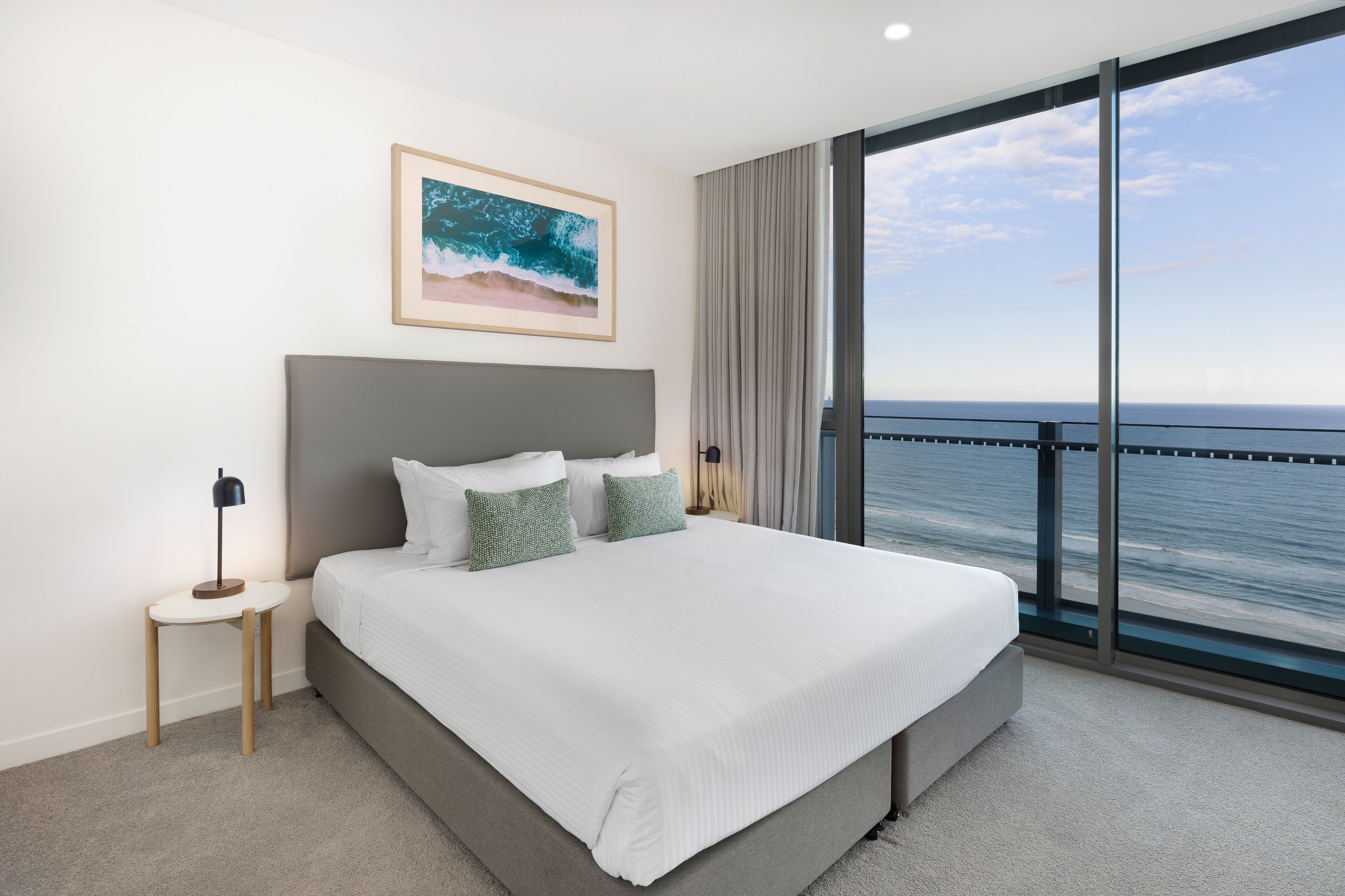 Una habitación de hotel moderna con una cama grande, una mesita de noche pequeña y un ventanal que ofrece vistas al océano.