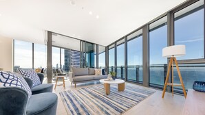 A modern living room with a sofa, coffee table, and floor lamp, large windows with a city view, and a balcony with a railing. - ULTIQA Signature at Broadbeach (Broadbeach)