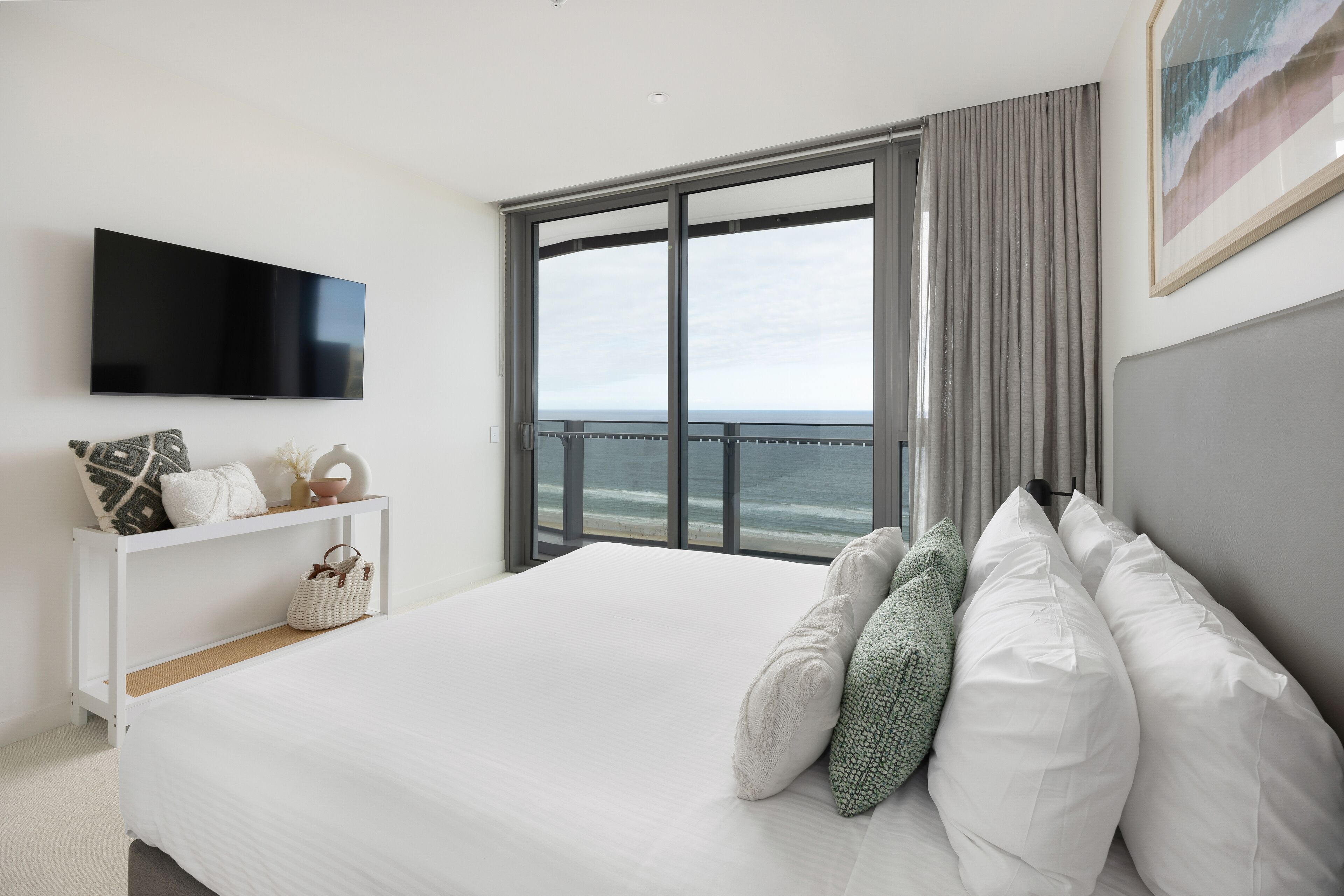 A modern hotel room with a large bed, a flat-screen TV, a view of the ocean, and a balcony with curtains.