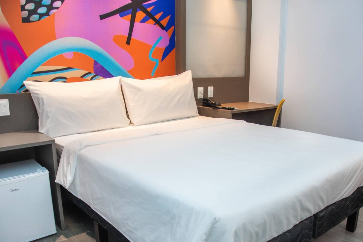 A hotel room with a bed, a nightstand, a desk, a chair, and a small refrigerator.