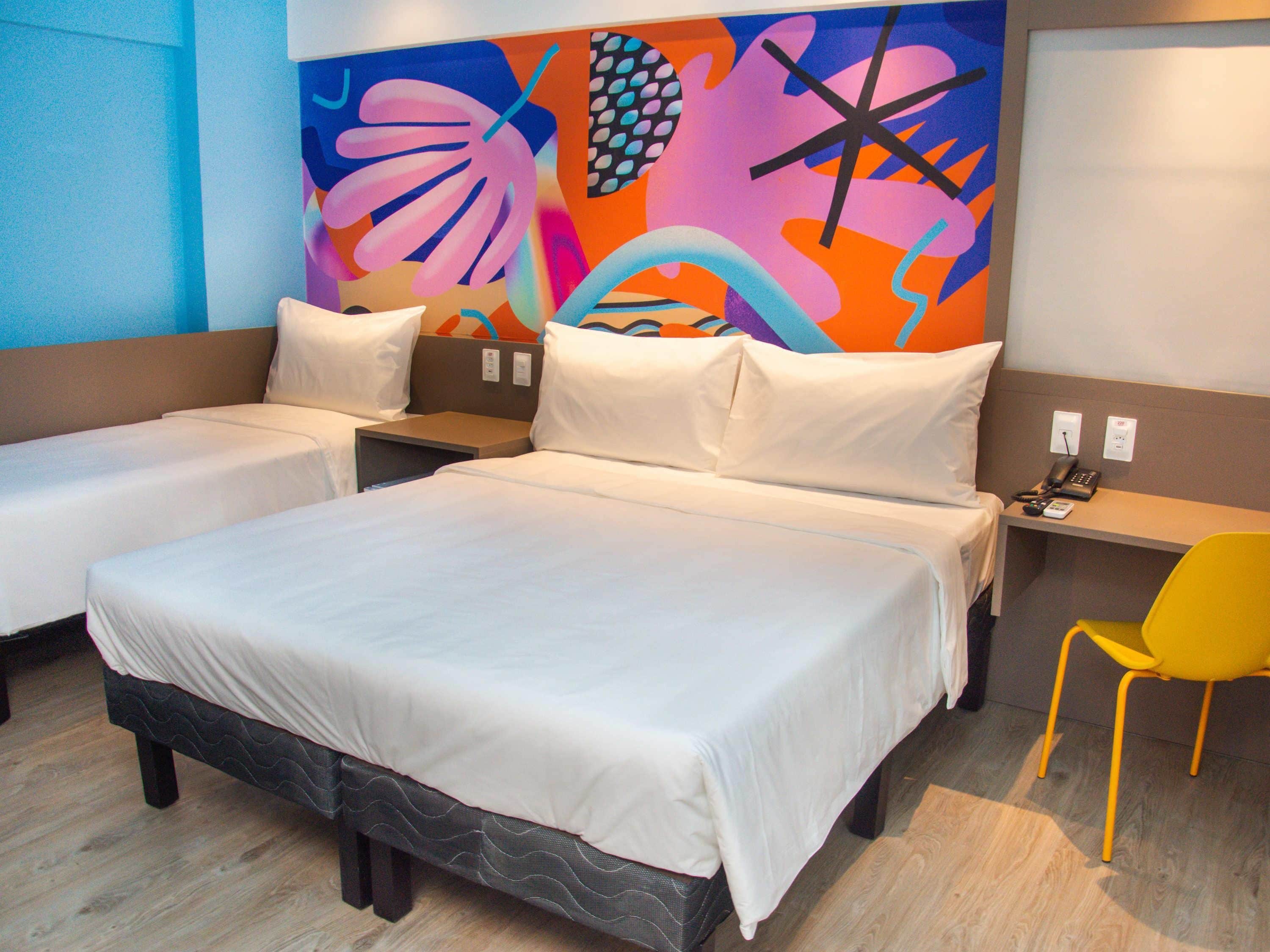 A hotel room with two single beds, a large bed, a desk with a chair, and a colorful abstract wall art.