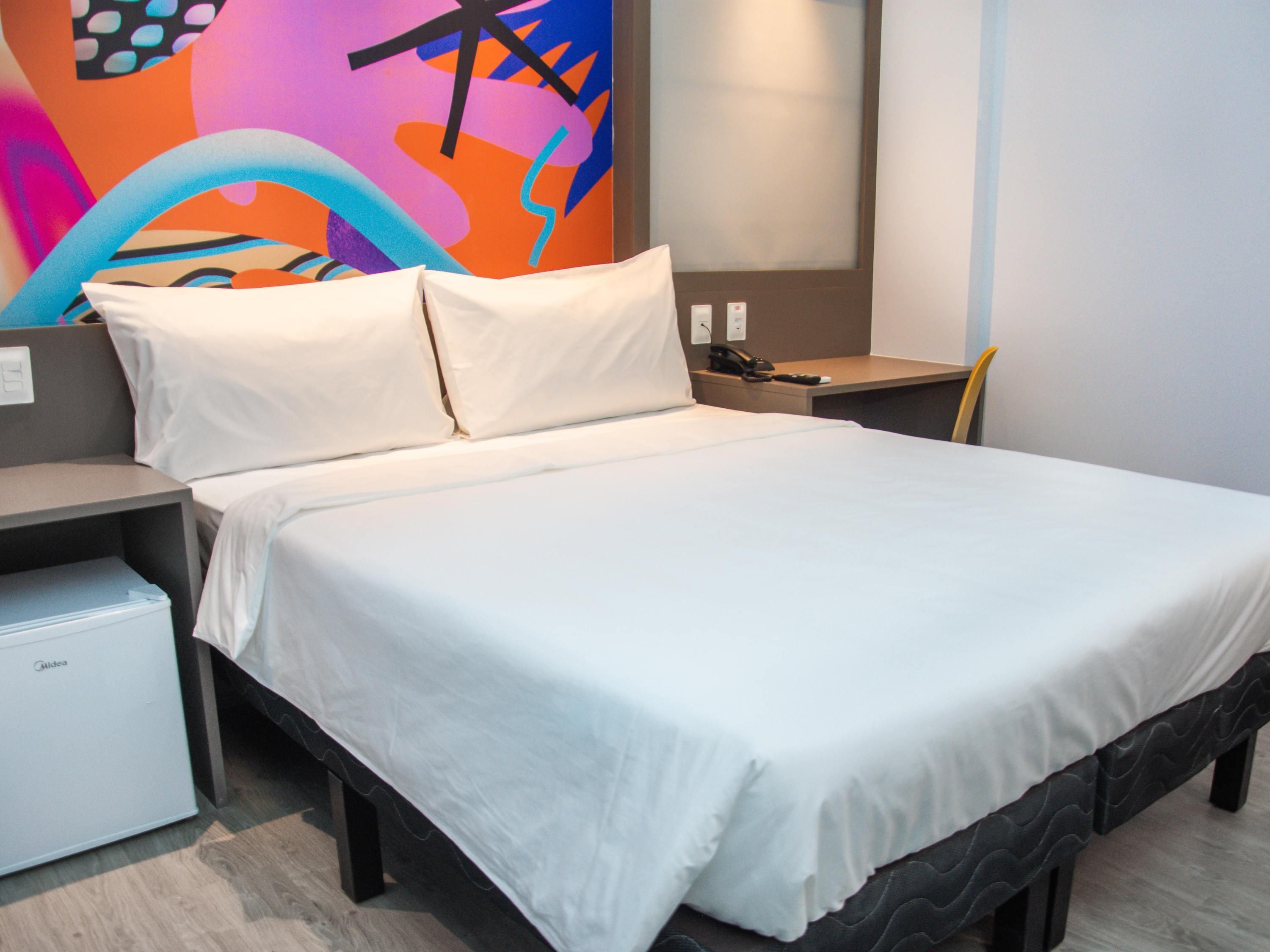 Quarto de hotel com cama, mesa de cabeceira, escrivaninha, cadeira e um pequeno frigorífico.