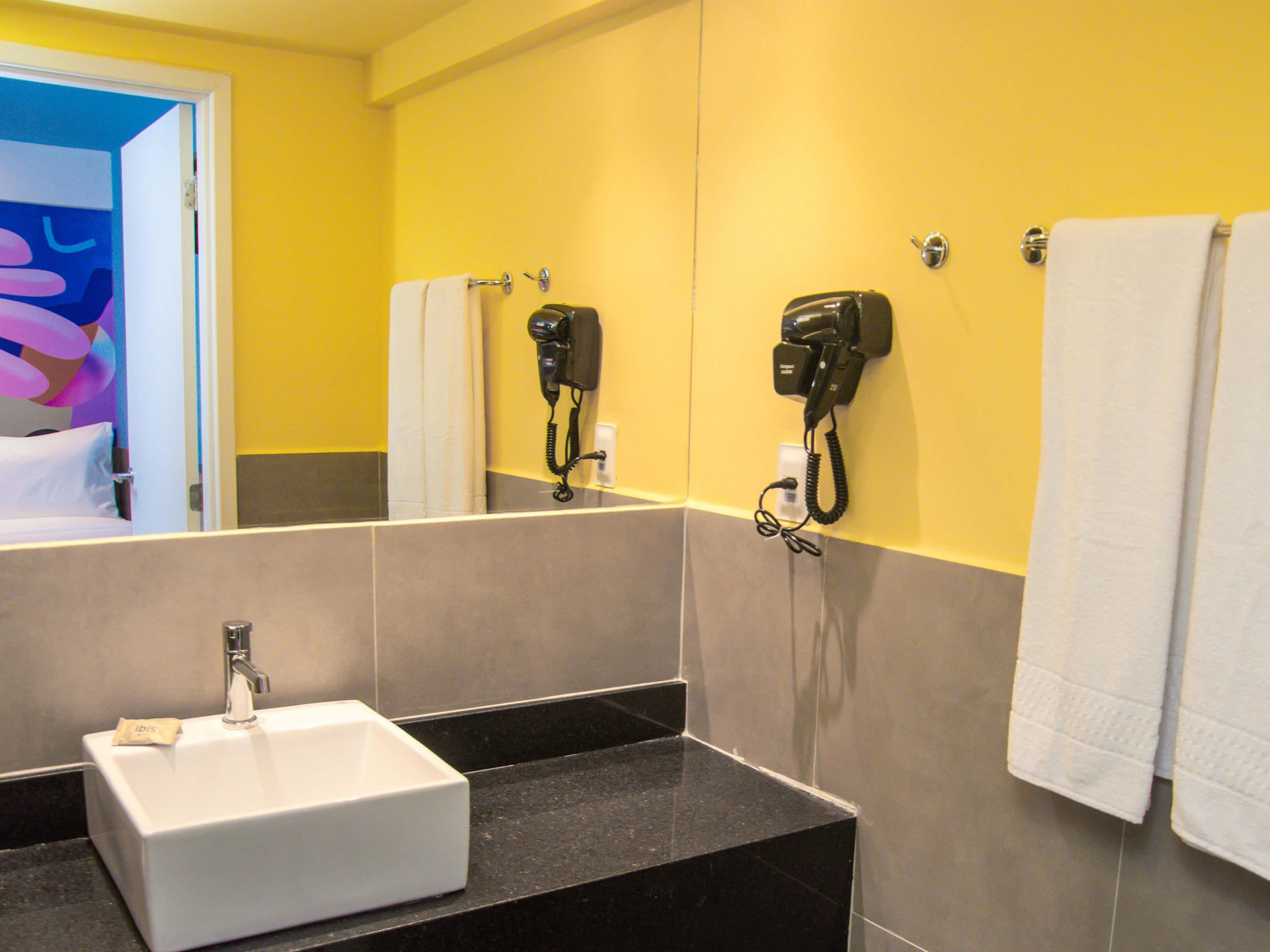 Un baño con un lavabo blanco, dos secadores de pelo fijados en la pared y un perchero con dos toallas.