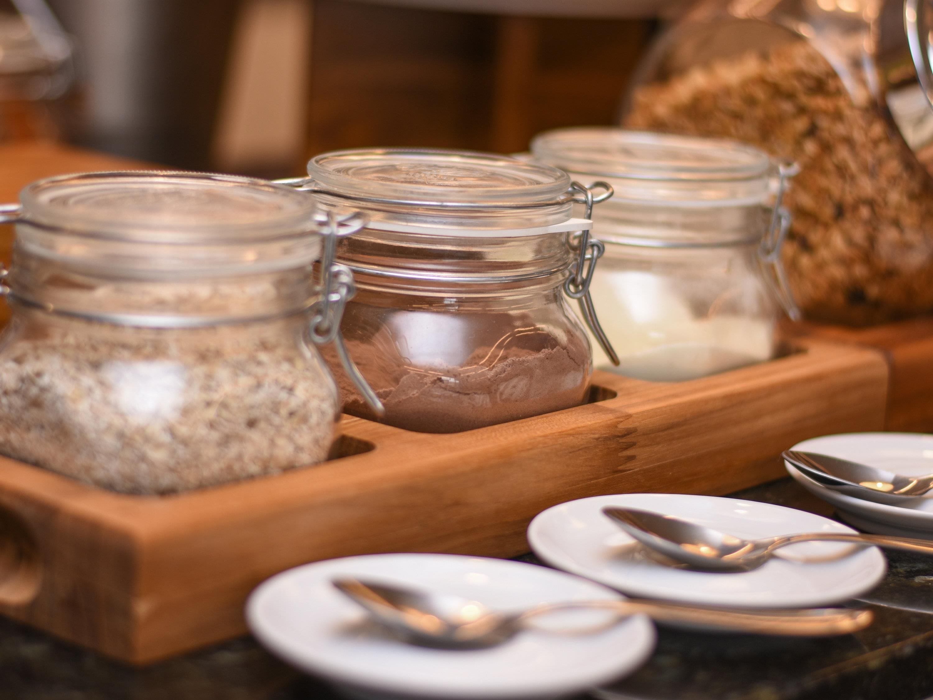 A breakfast buffet setup with glass jars containing various food items, a wooden serving tray, and white plates with silverware.