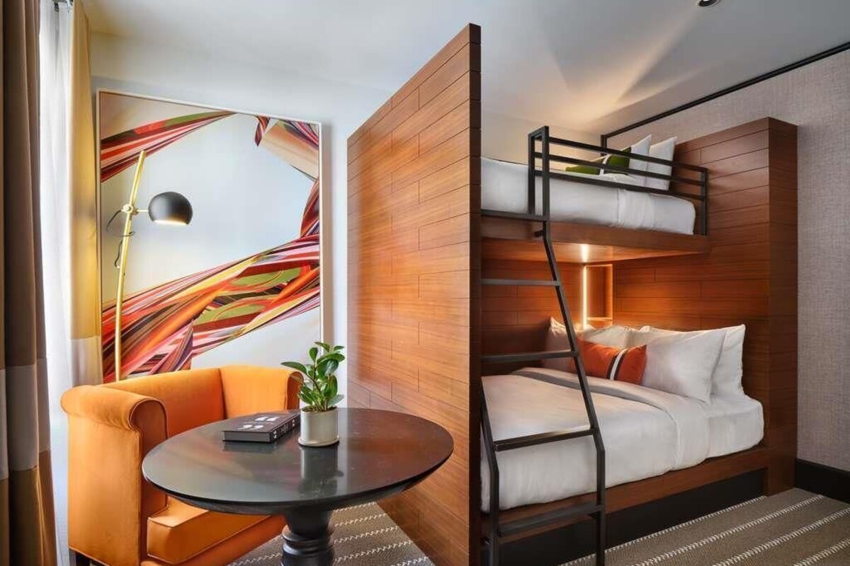 A modern hotel room with a bunk bed, a round table with a plant, an orange armchair, and a colorful abstract painting on the wall.