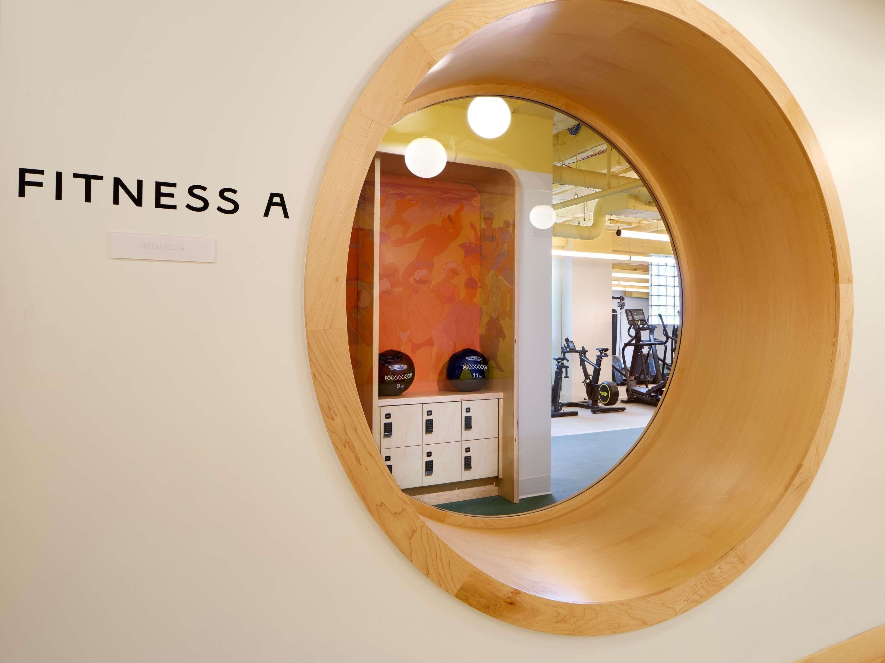 A circular wooden frame with a mirror reflecting a fitness area, including exercise equipment and a colorful wall.
