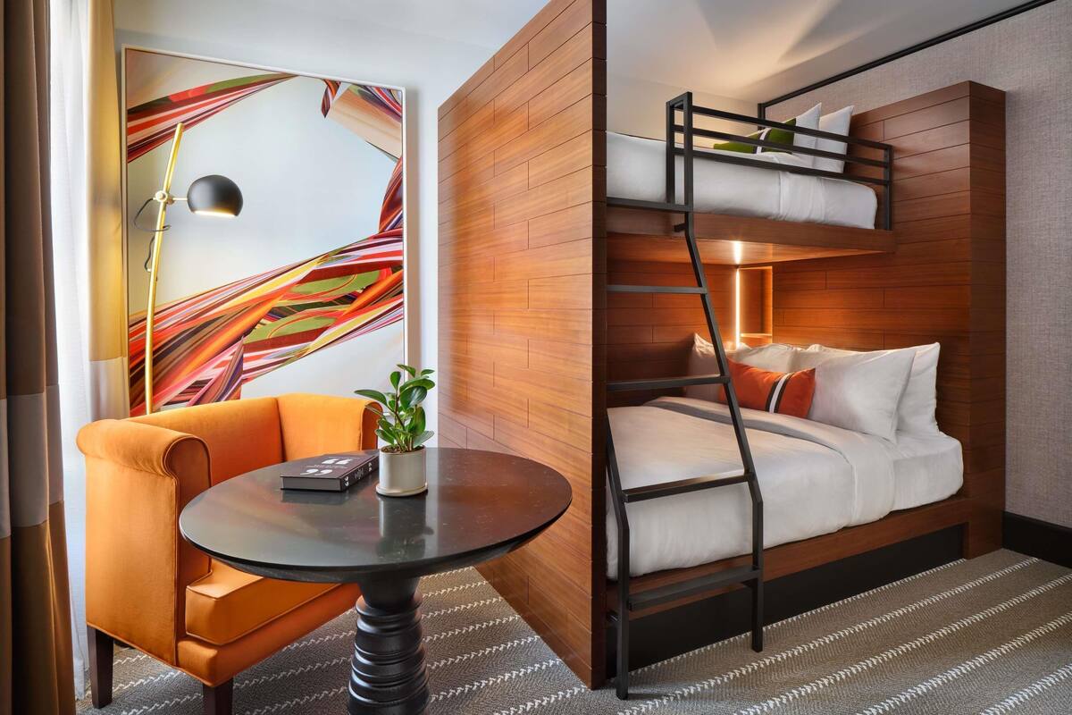 A hotel room with a bunk bed, a round table with a chair, a potted plant, and a colorful abstract painting on the wall.