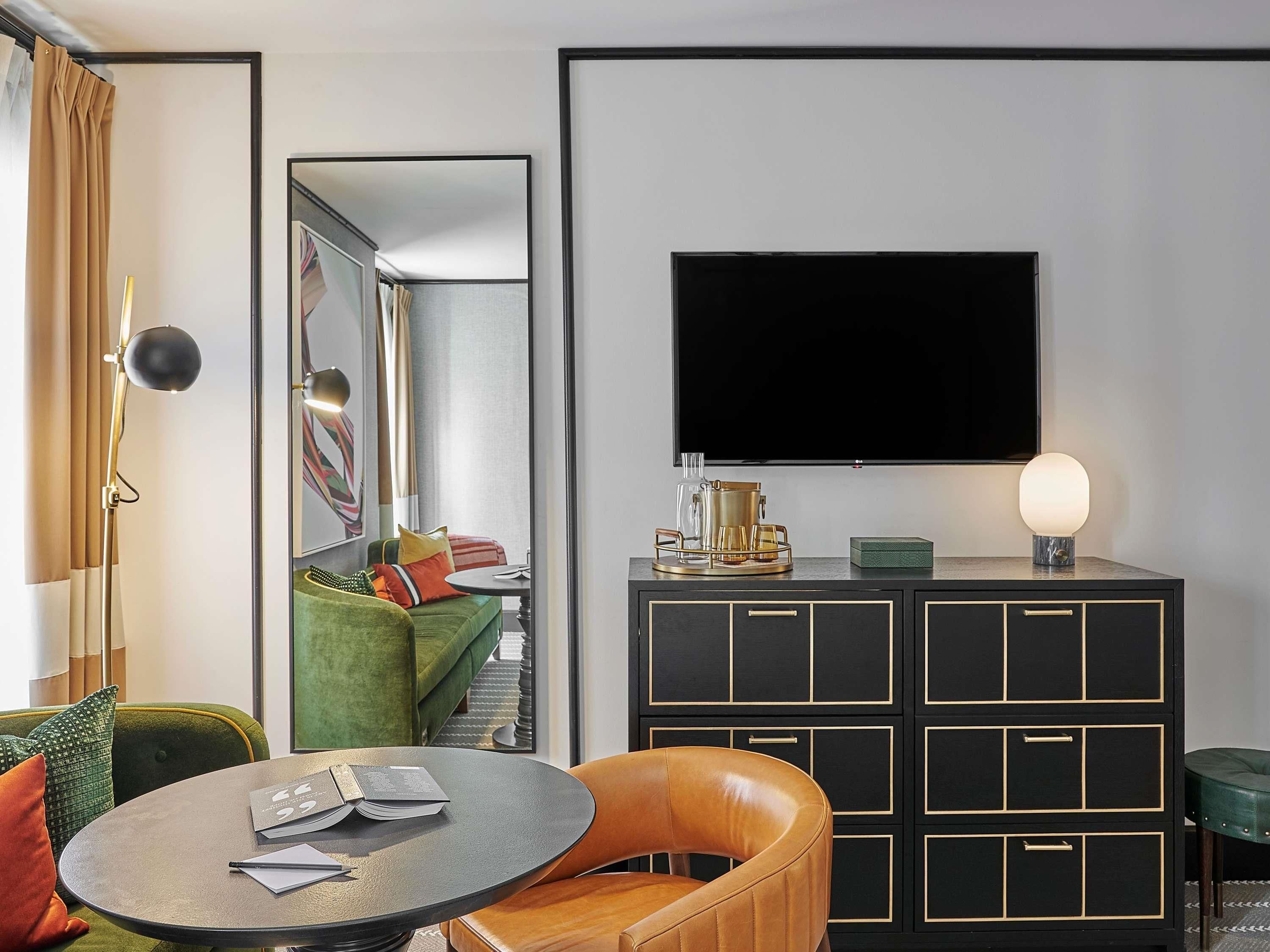 A modern hotel room with a flat-screen TV, a round table with a chair, a green sofa, and a black cabinet with drawers.