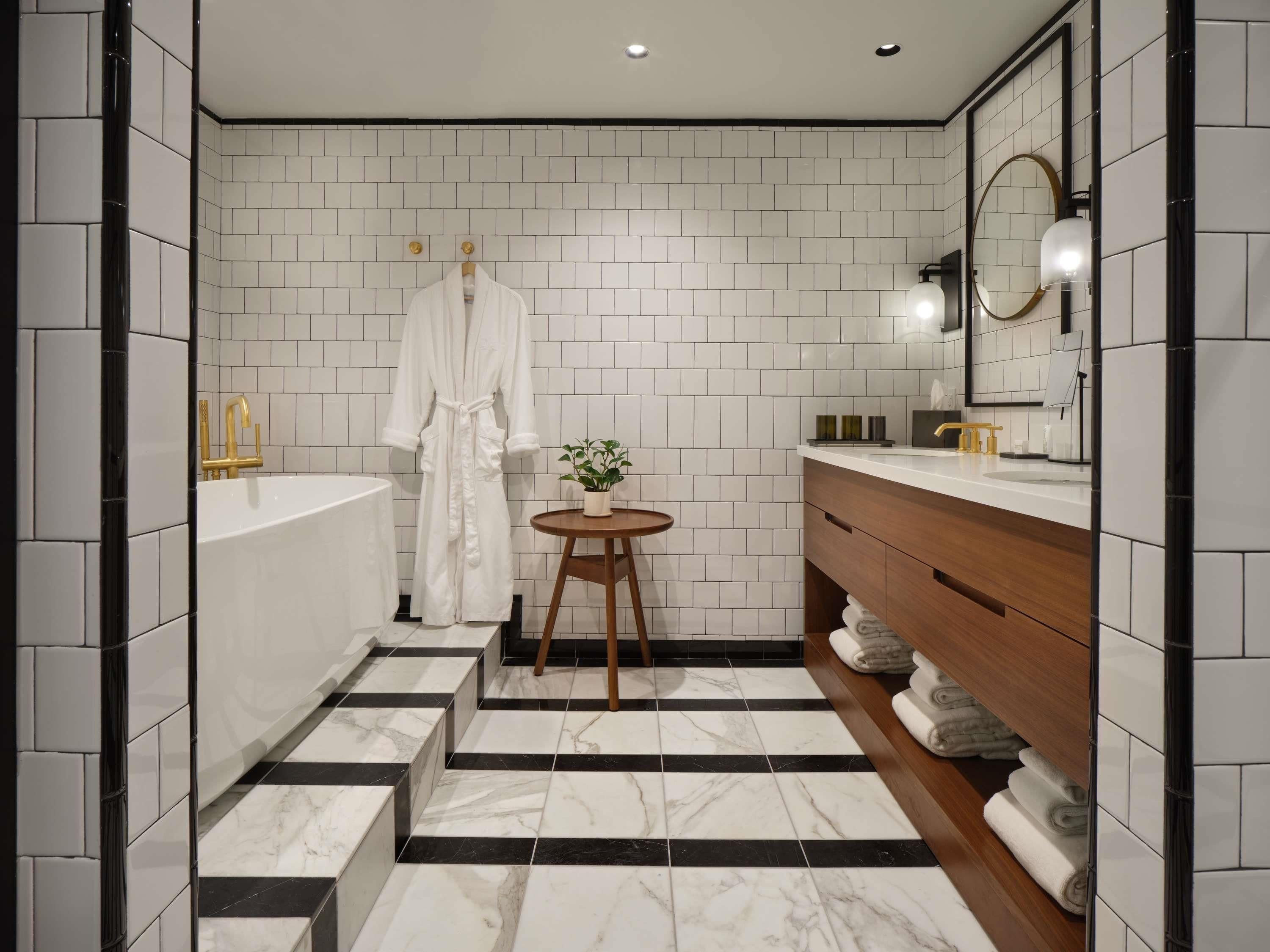 A modern bathroom with white subway tiles, a marble floor, a white bathtub, a wooden vanity, and a round wooden stool with a plant.