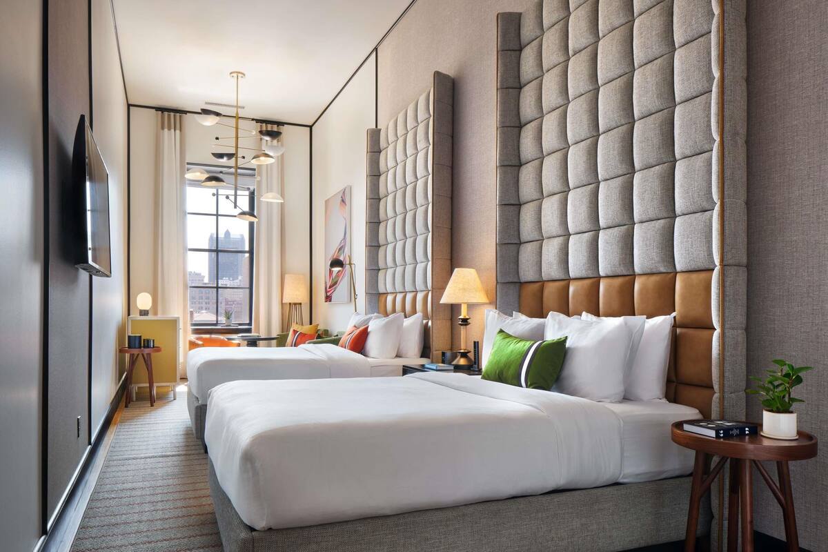 A hotel room with two beds, a large padded headboard, a flat-screen TV, a window with a city view, and a small side table with a plant.