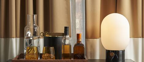Una bandeja de madera con botellas y vasos sobre un mueble oscuro con pomos dorados, frente a cortinas color beige.