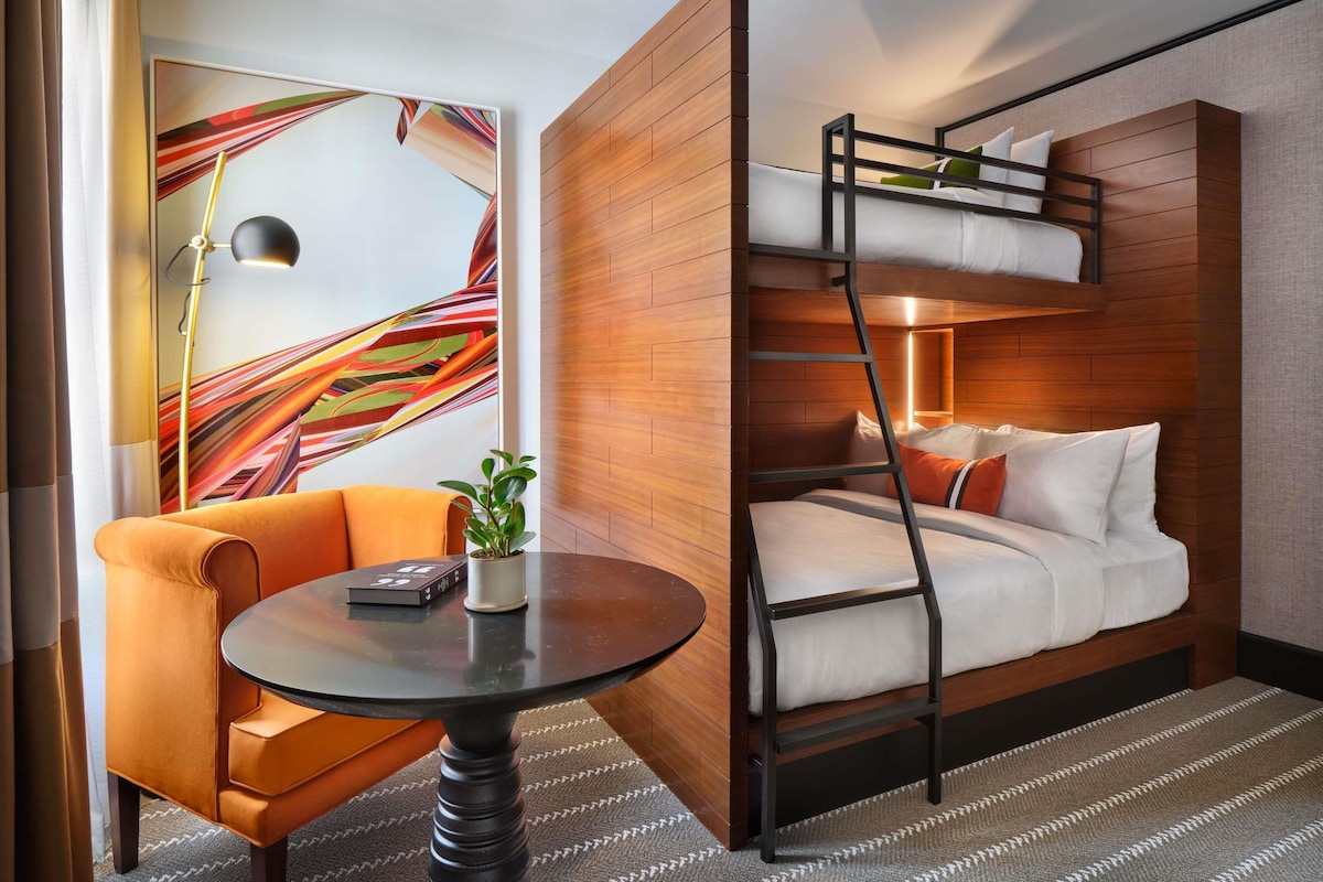 A hotel room with a bunk bed, a round table with a chair, a potted plant, and a colorful abstract painting on the wall.