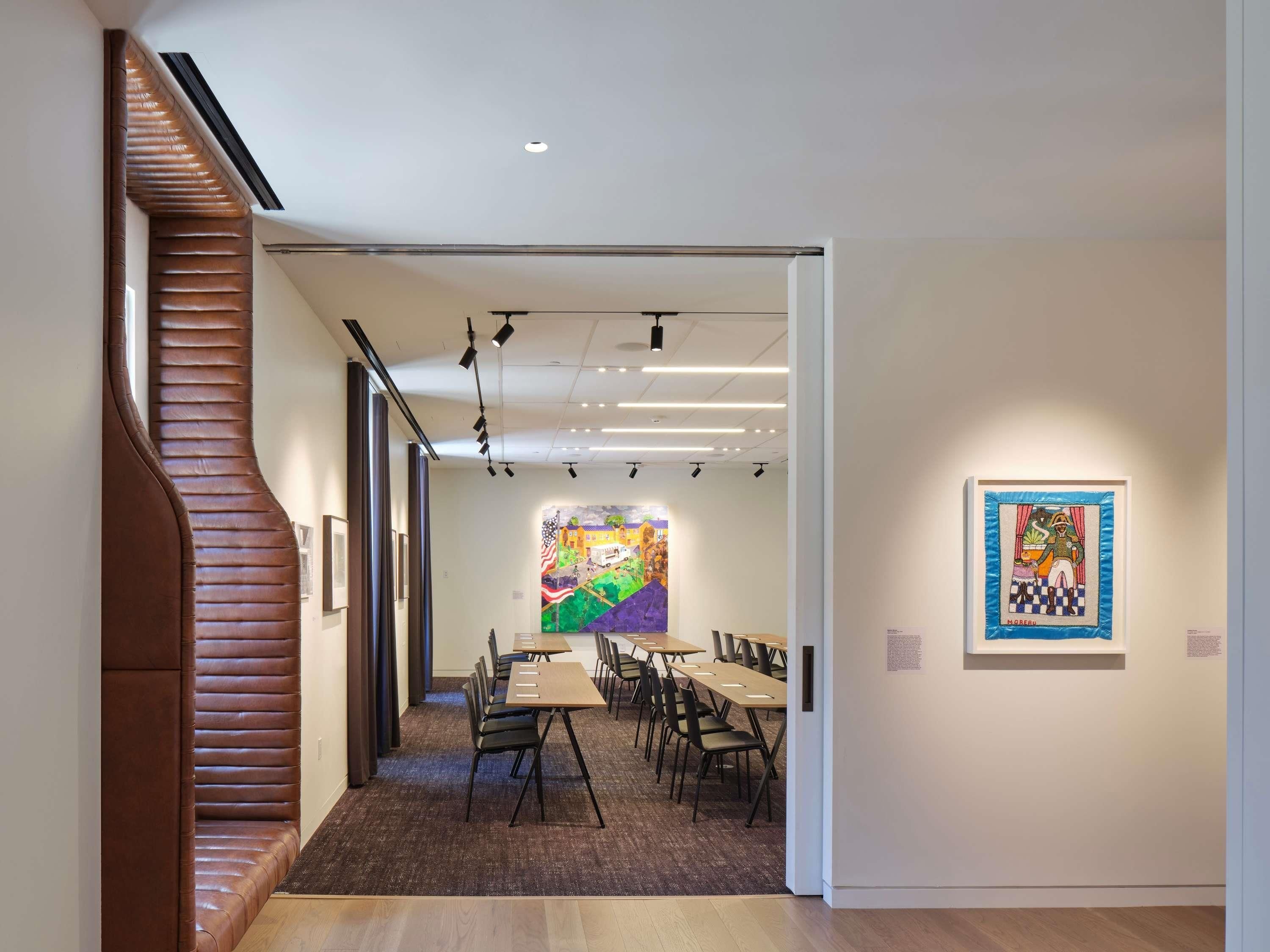Une galerie d’art dotée d’un intérieur moderne, avec une table centrale entourée de chaises, une fresque murale colorée et des œuvres d’art encadrées accrochées aux murs.