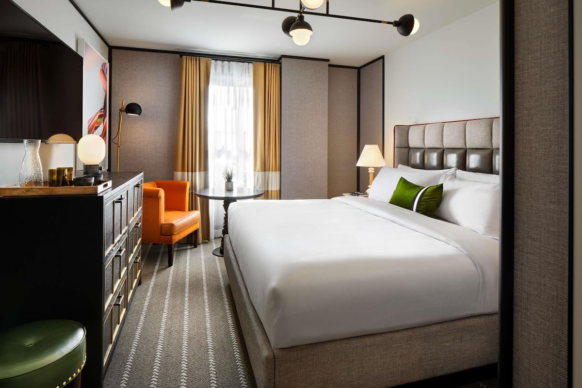 A hotel room with a large bed, a desk with a chair, a small table, and a lamp.