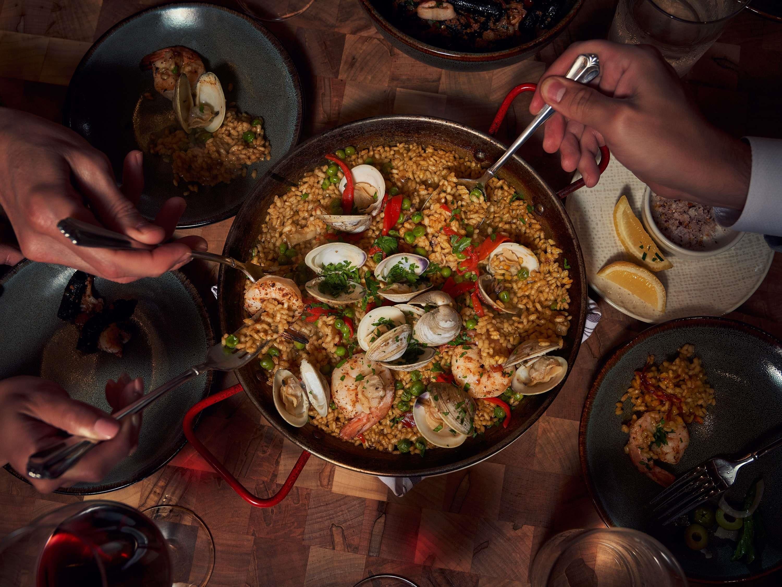 A large pan of seafood paella with clams and shrimp, surrounded by diners.