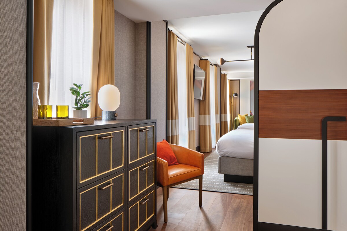 A hotel room with a wooden floor, a desk with drawers, a chair, a bed with a headboard, and curtains.