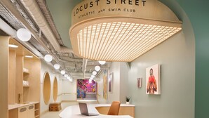 Un área de recepción con un escritorio de madera curvada, una computadora portátil y una silla. En la pared, un letrero que dice “Locust Street Athletic and Swim Club”. 