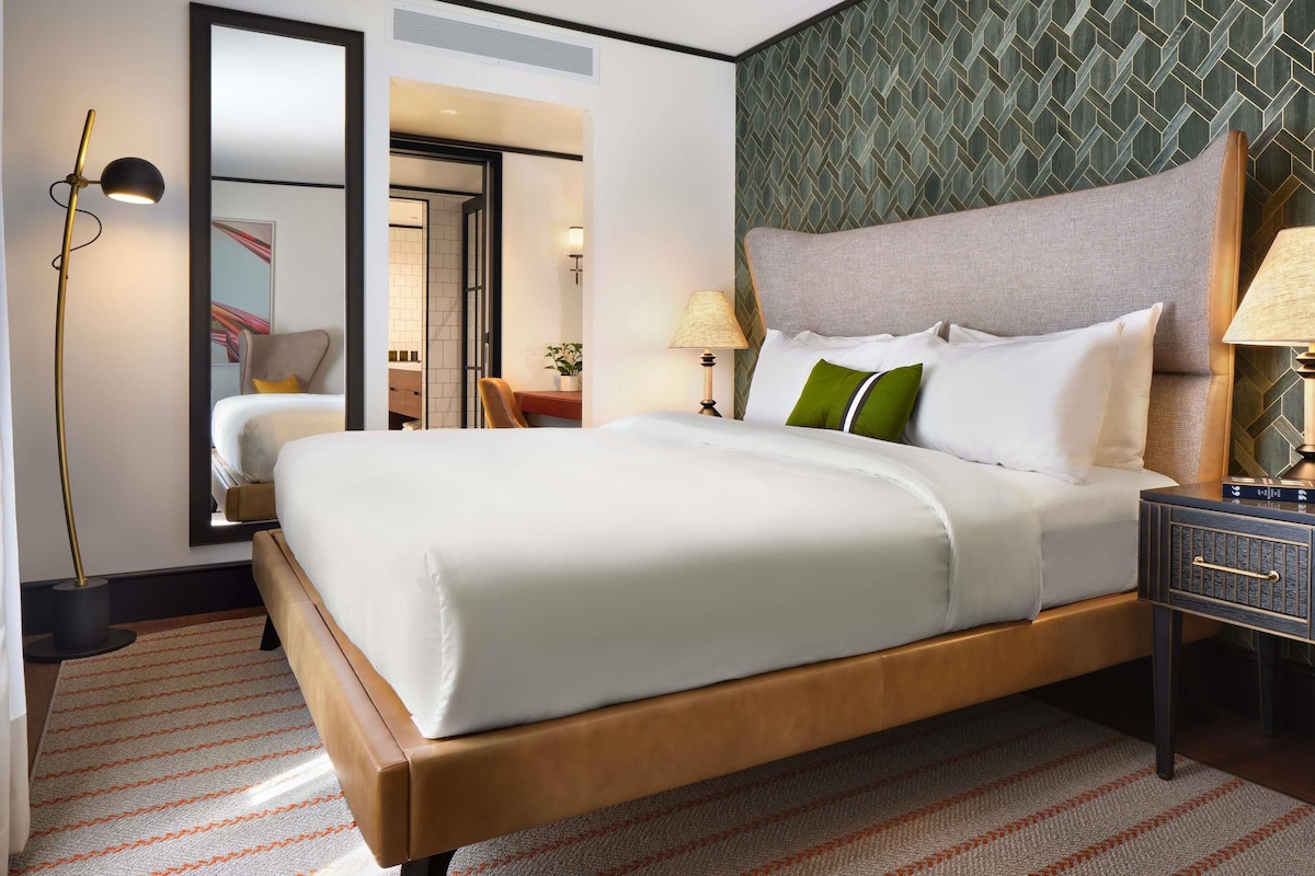 A hotel room with a large bed, a desk, a chair, a nightstand, and a mirror.