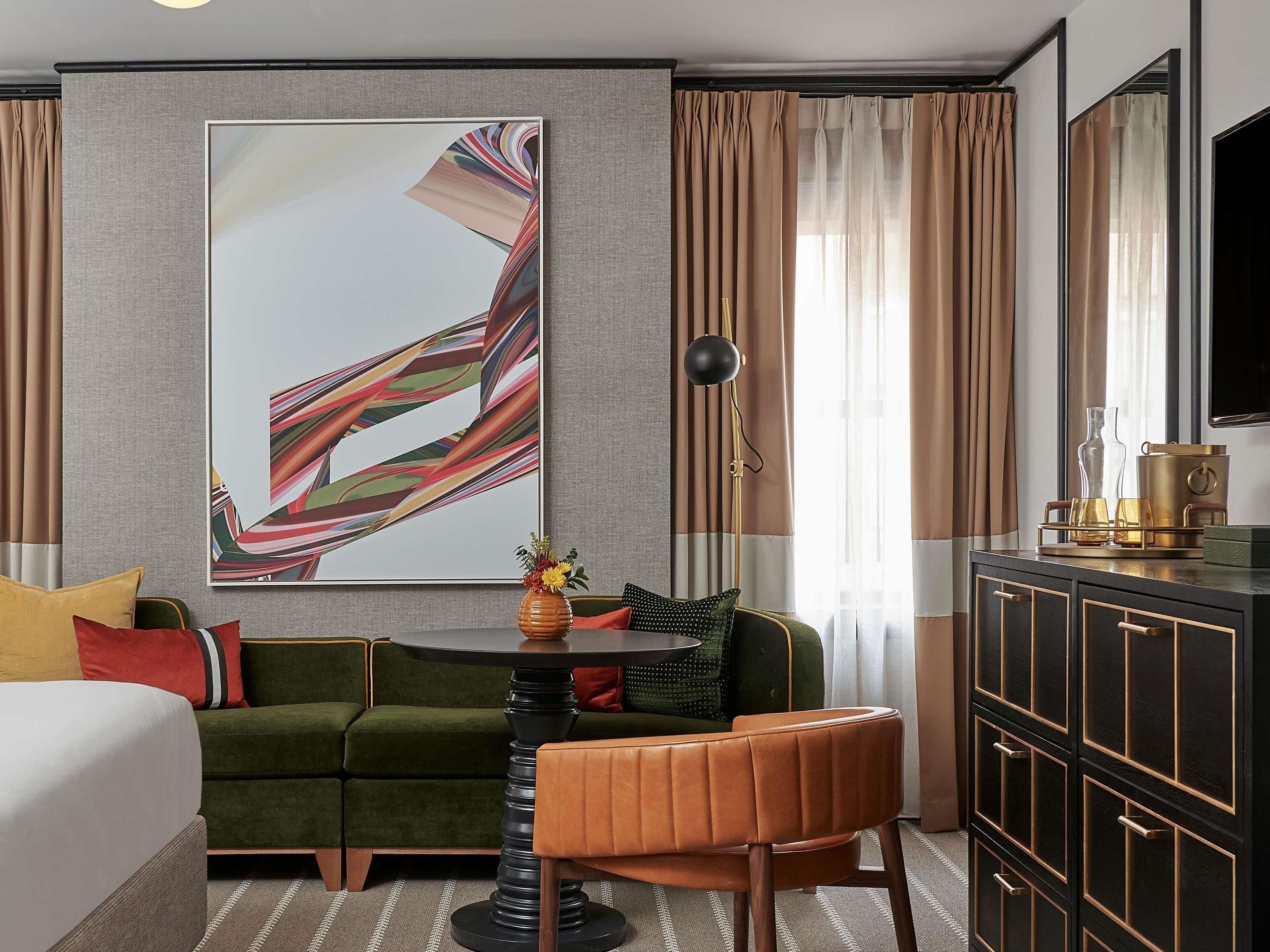 A hotel room with a green sofa, a small round table, a single chair, a bed, a large abstract painting, a floor lamp, a cabinet with a mirror, and curtains.