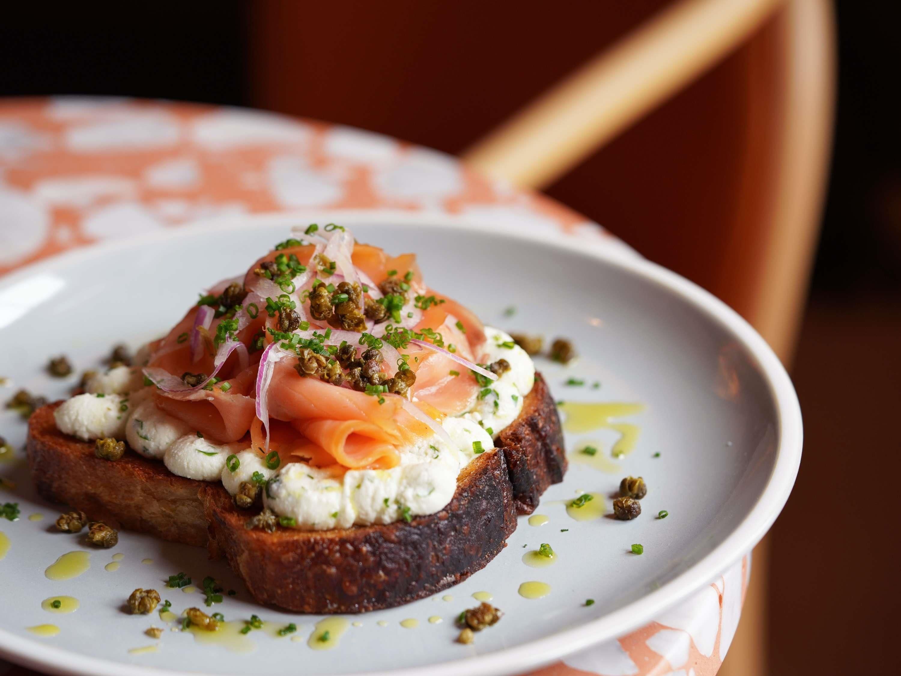 Un plato con un sándwich cubierto de salmón ahumado, queso crema y alcaparras, decorado con cebollino.