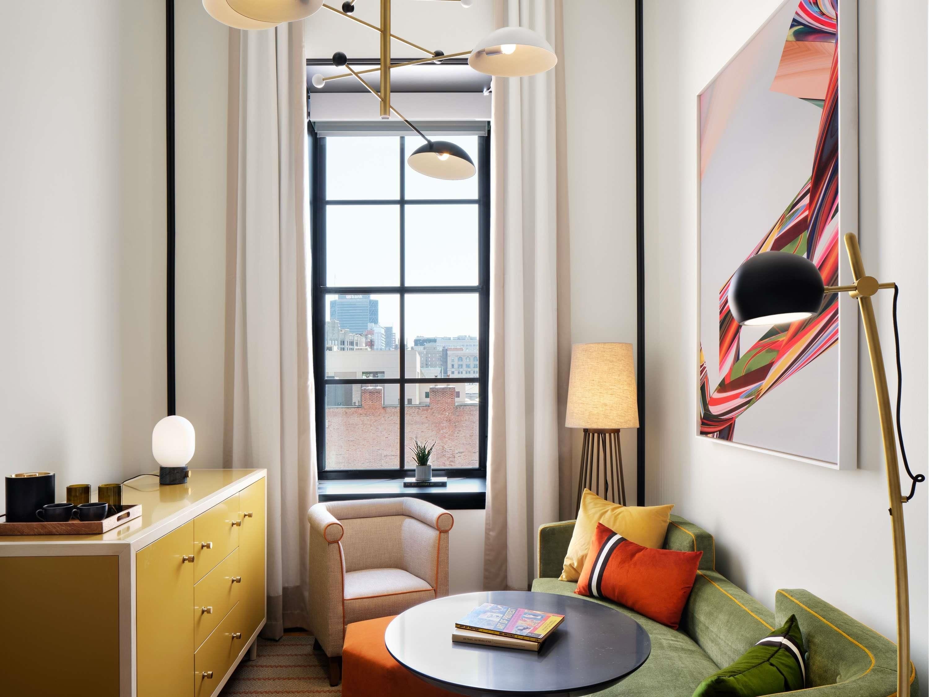 A modern living room with a sofa, a small table, a chair, a window with a view of buildings, and a lamp.