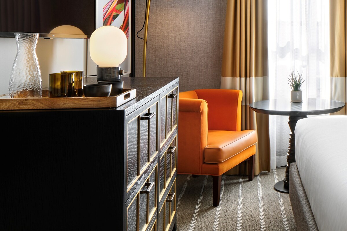 A hotel room with a bed, an orange armchair, a small round table, a desk with a lamp, and a decorative vase.