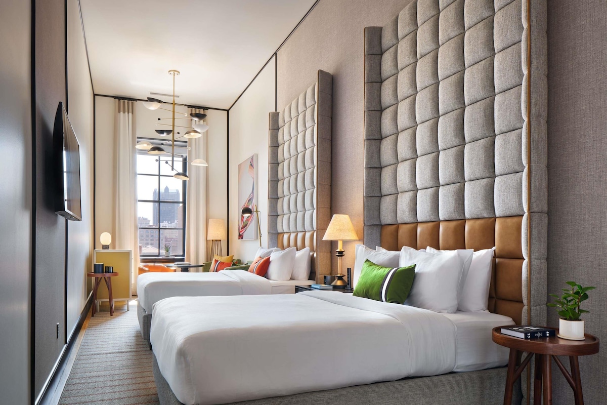 A hotel room with two beds, a large padded headboard, a flat-screen TV, a window with a city view, and a small side table with a plant.