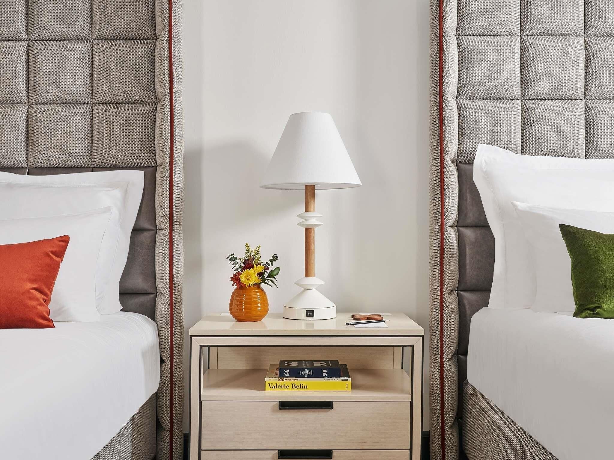 Une chambre d’hôtel avec deux lits, une table de chevet avec une lampe, un vase avec des fleurs et un livre.