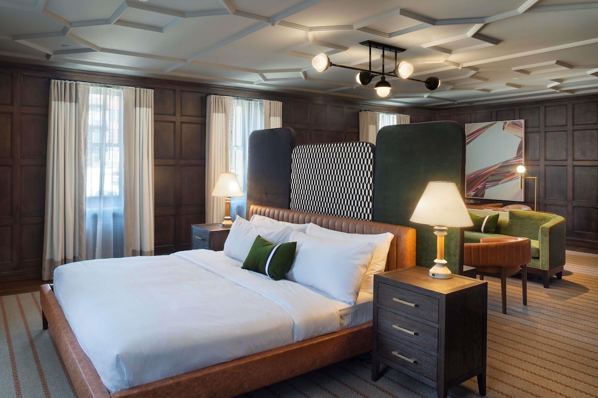A hotel room with a large bed, a wooden headboard, a nightstand with a lamp, a green armchair, and a large painting on the wall.