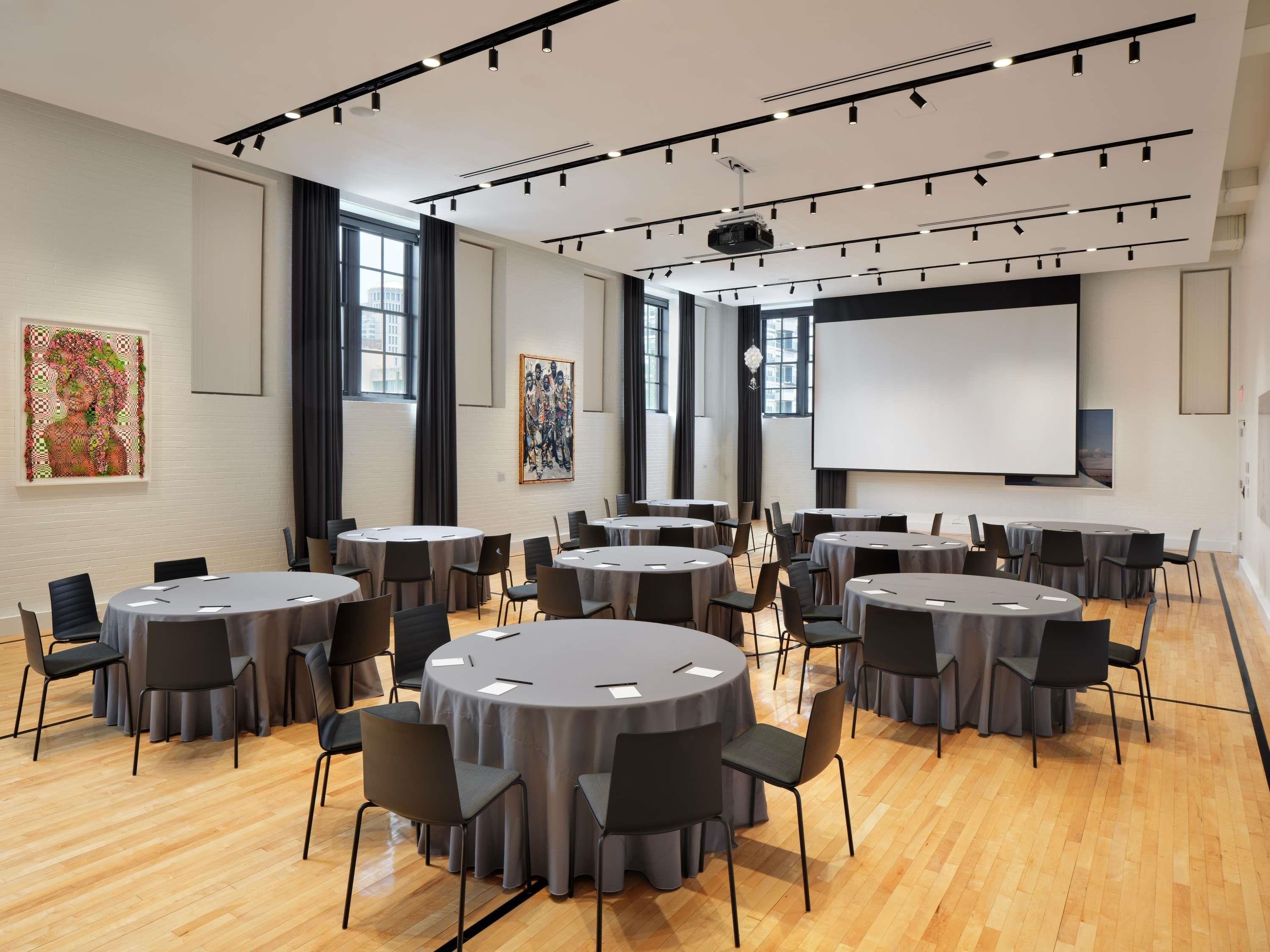 A conference room with multiple round tables, chairs, a projector screen, and artwork on the walls.