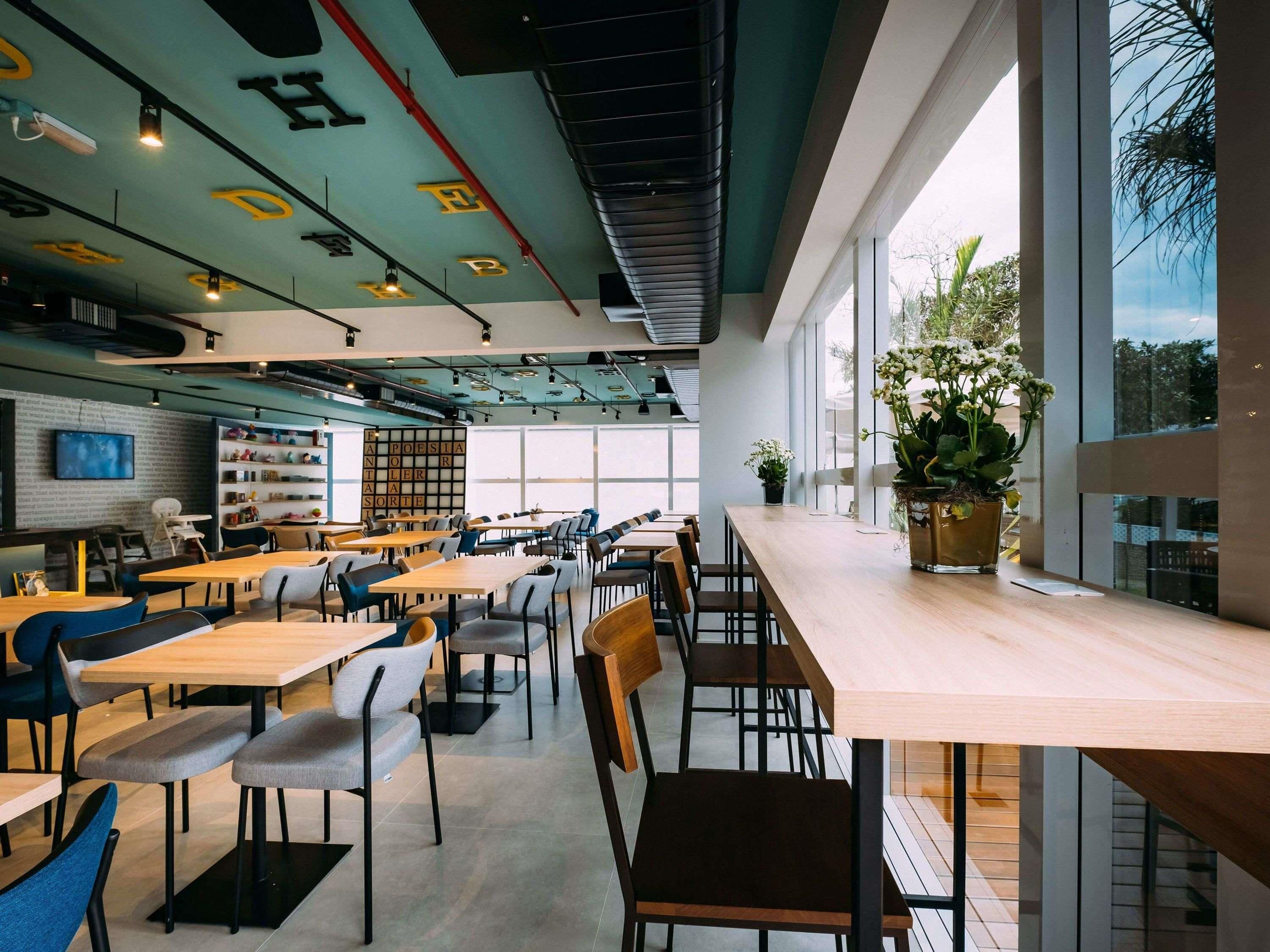 A modern cafe with multiple tables and chairs, a bookshelf, and large windows offering a view of the outdoors.