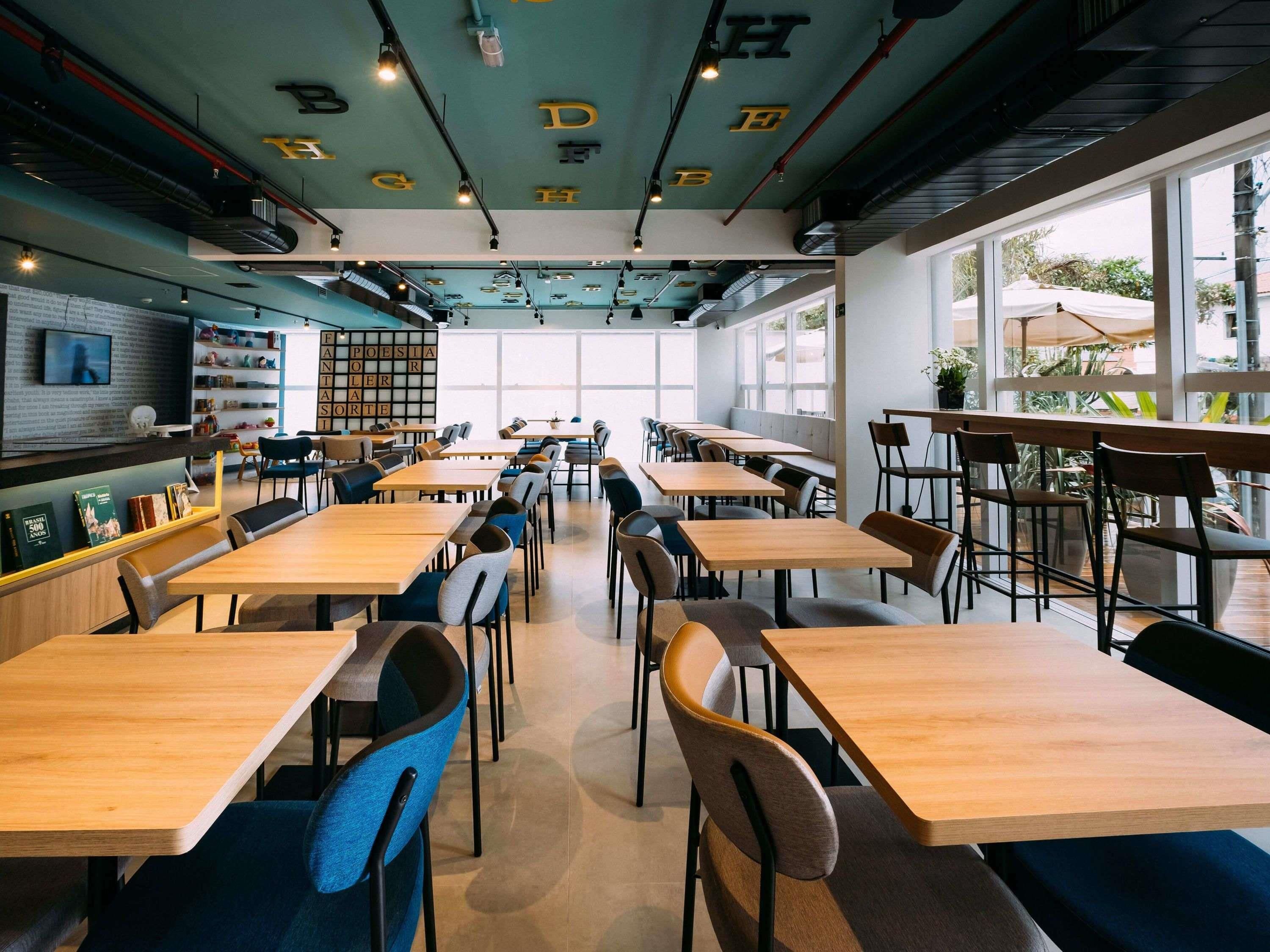Un interior de cafetería moderno con diversas opciones de asientos, una estantería visible y grandes ventanales que dejan entrar luz natural.