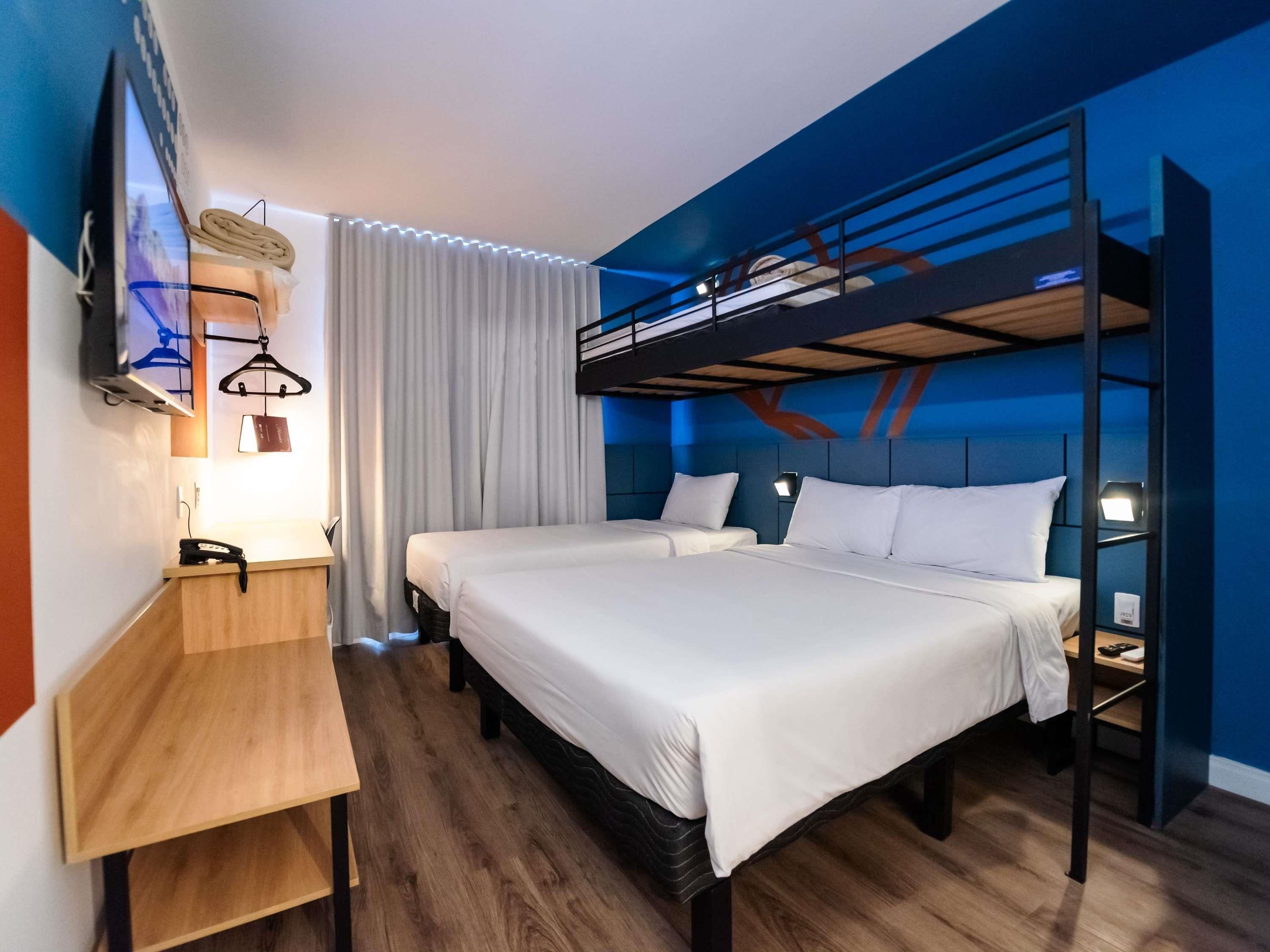 A hotel room with a bunk bed, a desk with a phone, a lamp, and a TV mounted on the wall.