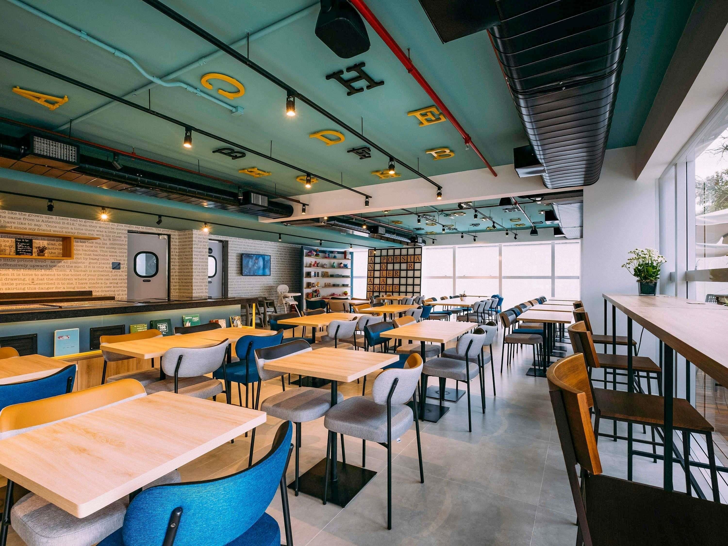 Un caffè moderno con diversi tavoli e sedie, una libreria ben visibile e un'ampia vetrata che fa entrare molta luce naturale.